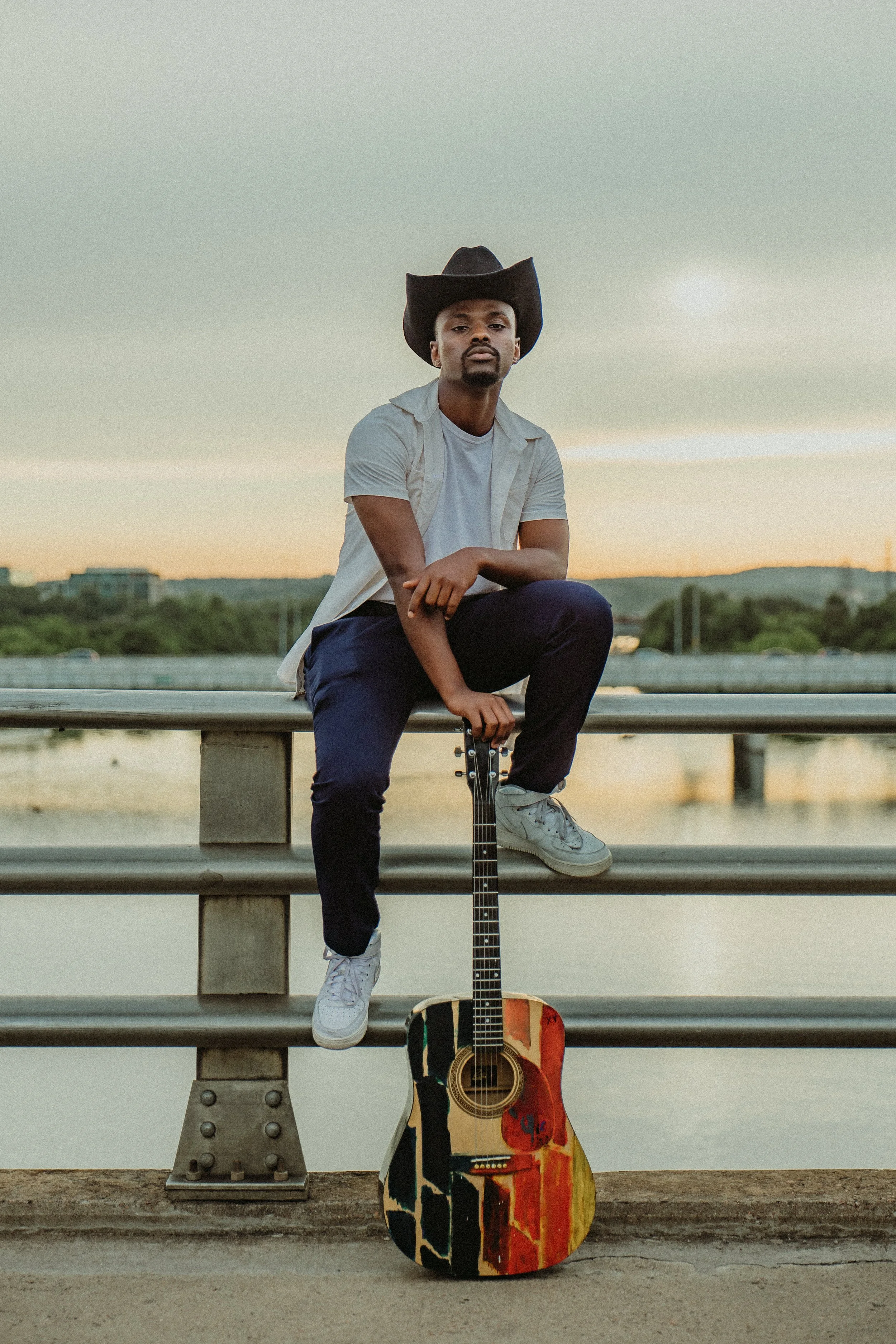 A man wearing a cowboy hat, white t-shirt, and dark pants sits on a bridge railing at sunset, holding a colorful acoustic guitar.