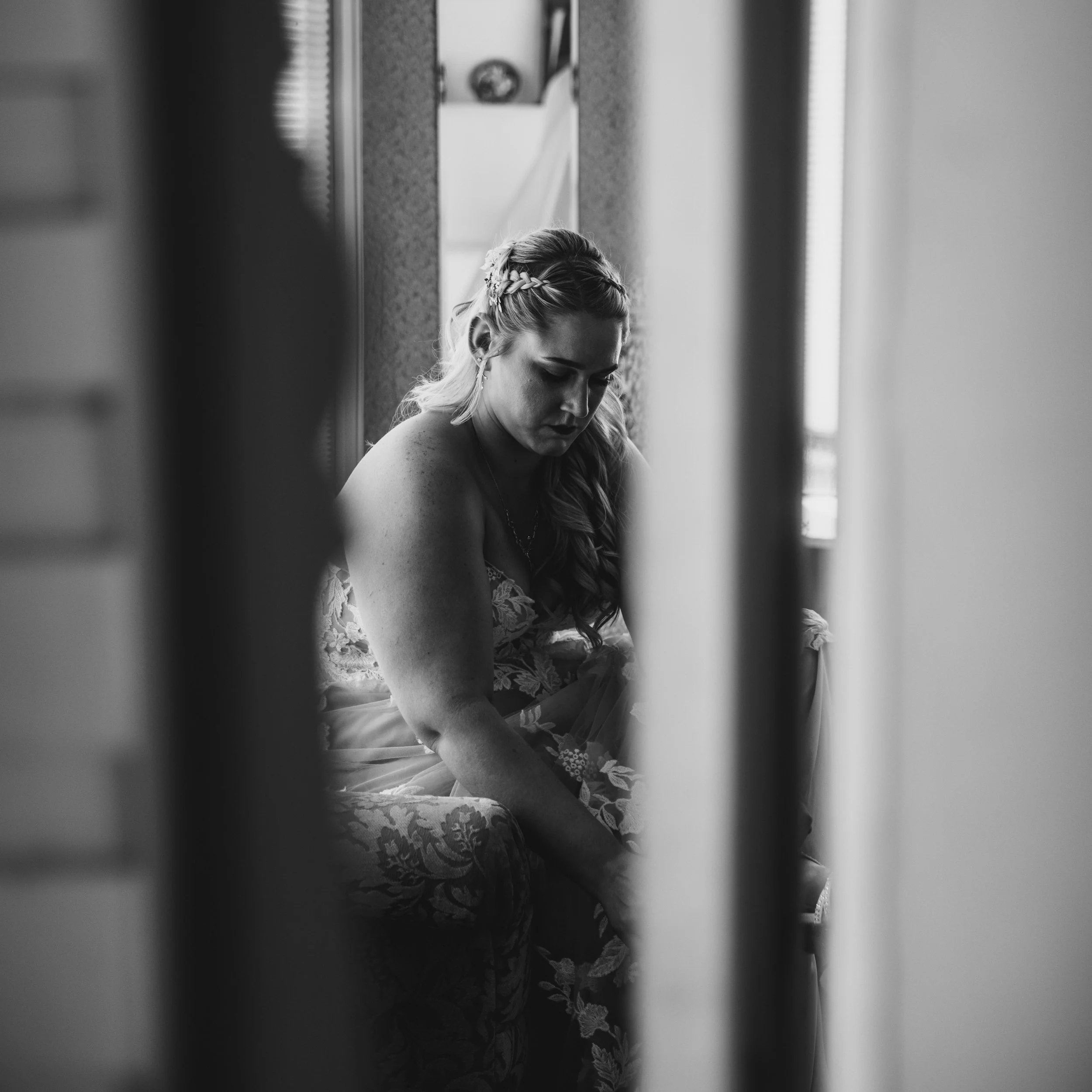 A woman in a lace dress sitting on a cushioned chair, seen through a narrow gap, possibly preparing for a special occasion.