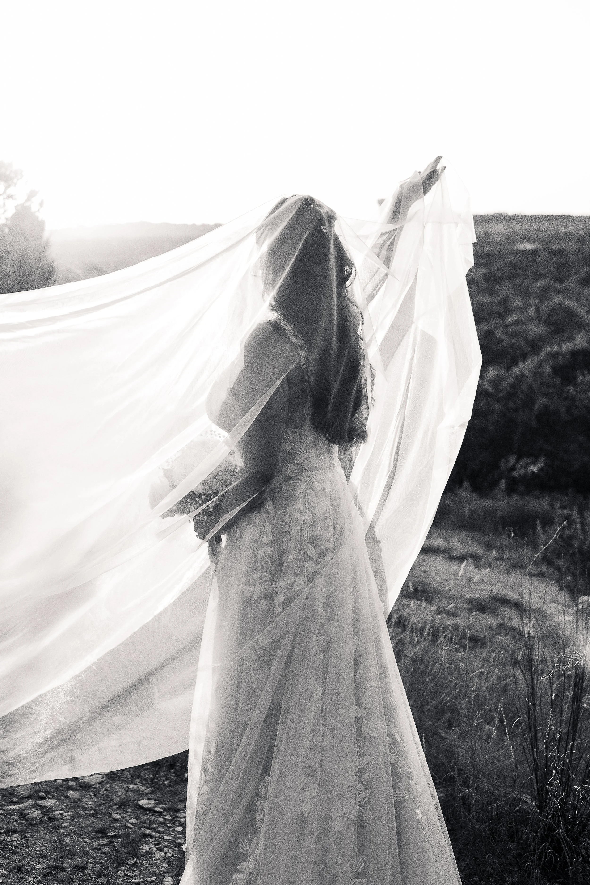 A woman wearing a wedding dress with floral details standing outdoors with a long veil flowing in the wind at sunset.