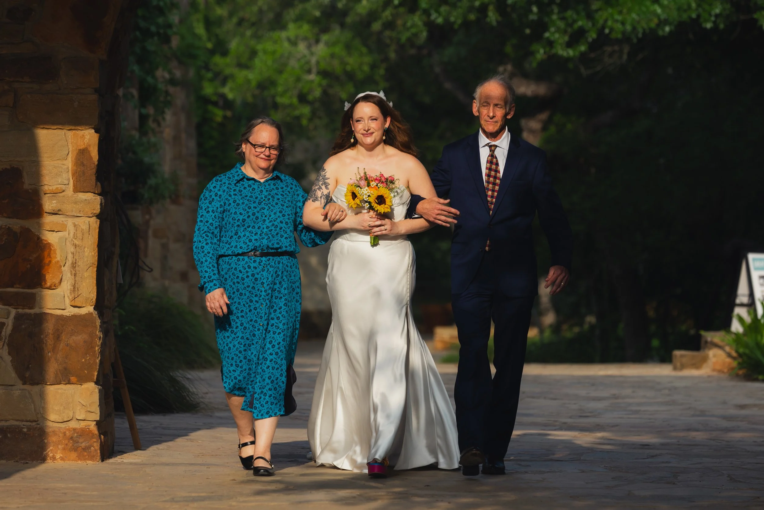 A bride in a white wedding gown holding a bouquet of sunflowers and pink flowers is walking down the aisle with her parents, navigating a stone pathway outdoors.