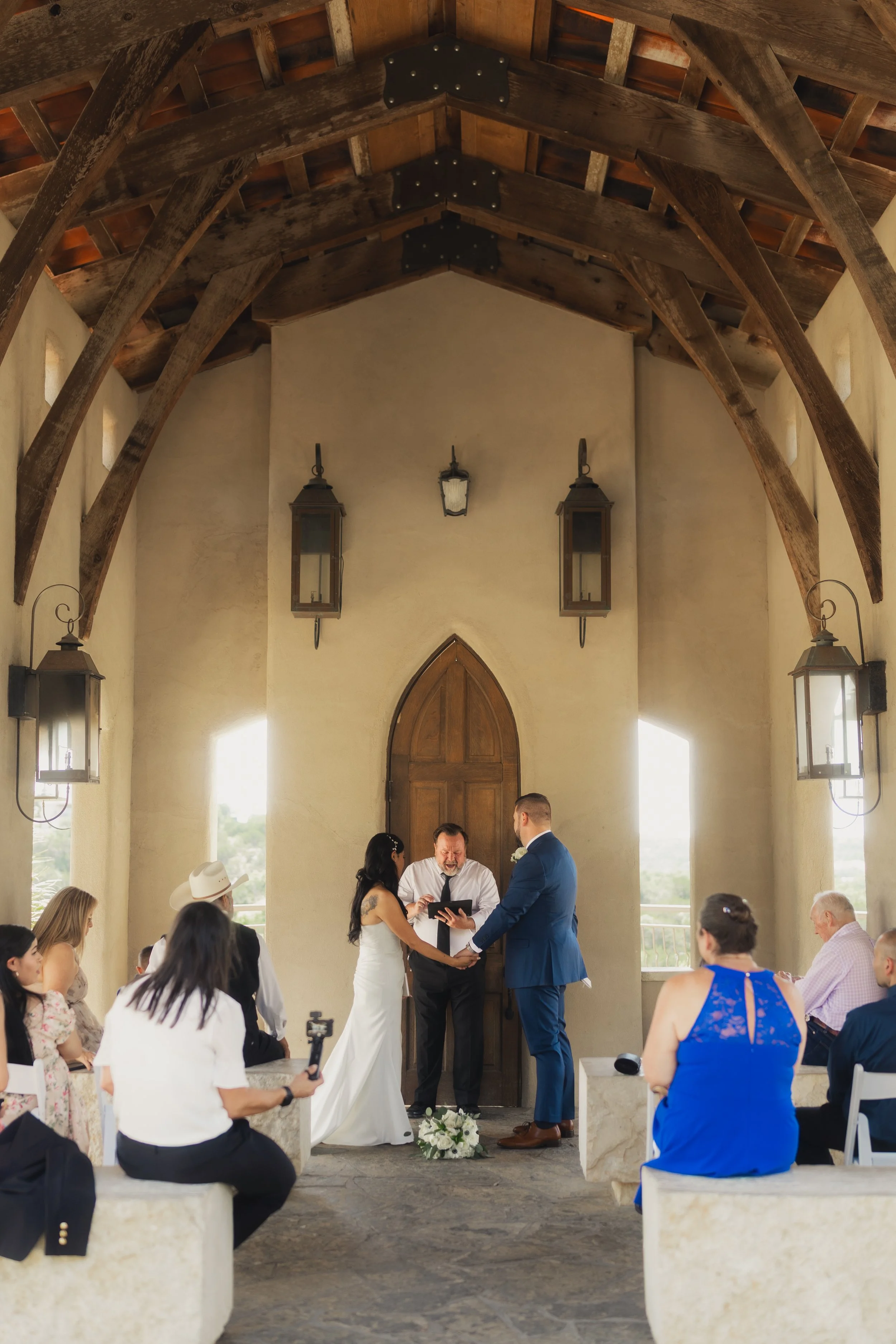 A couple gets married at an outdoor ceremony in a rustic setting with wooden beams. The bride and groom hold hands in front of an officiant, surrounded by seated guests. The scene is bright and airy, with natural light coming through open sides.