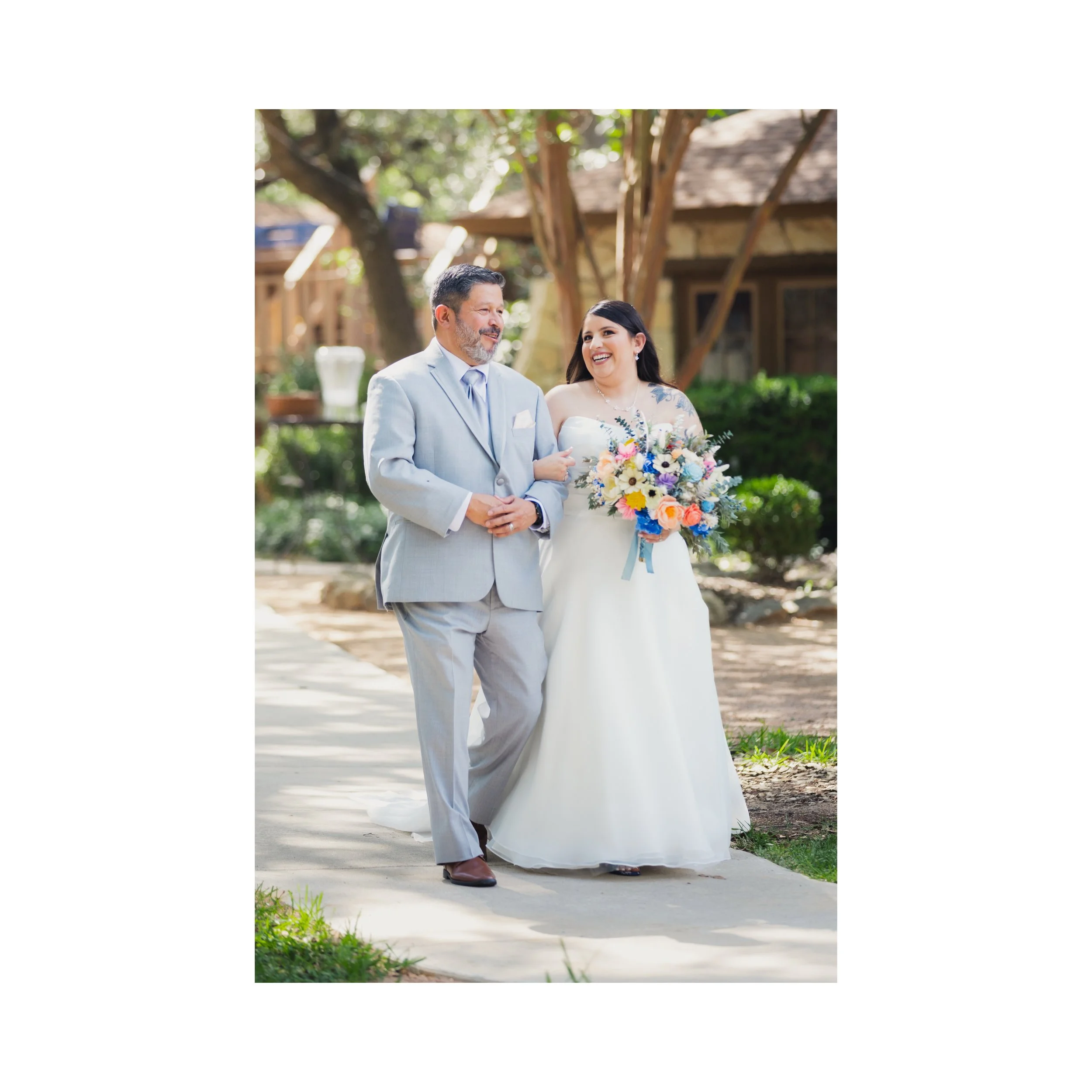 A bride in a white wedding dress holding a bouquet of colorful flowers walking outdoors with a man in a light gray suit, both smiling and looking at each other under a canopy of trees.