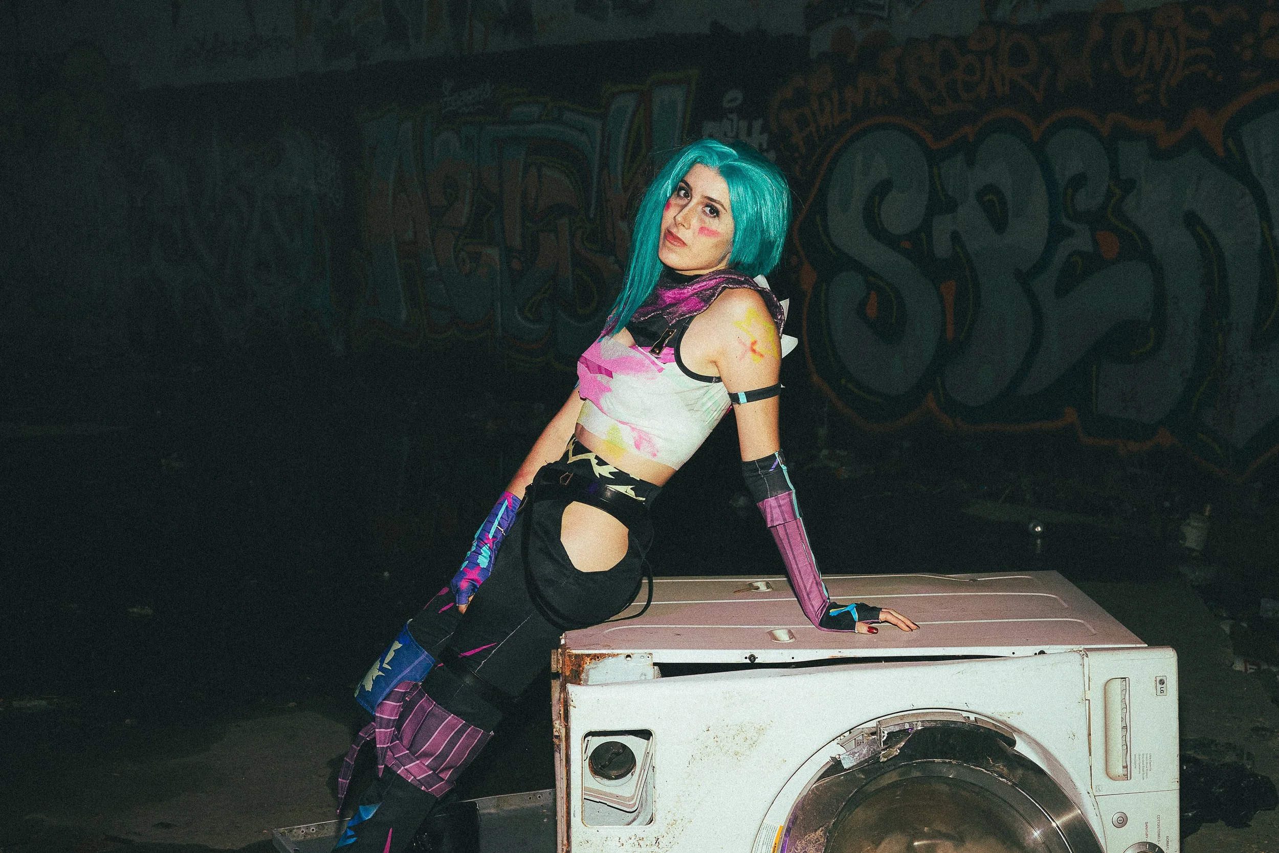 A woman with bright blue hair and colorful clothing leaning on a washing machine in a graffiti-covered urban setting.