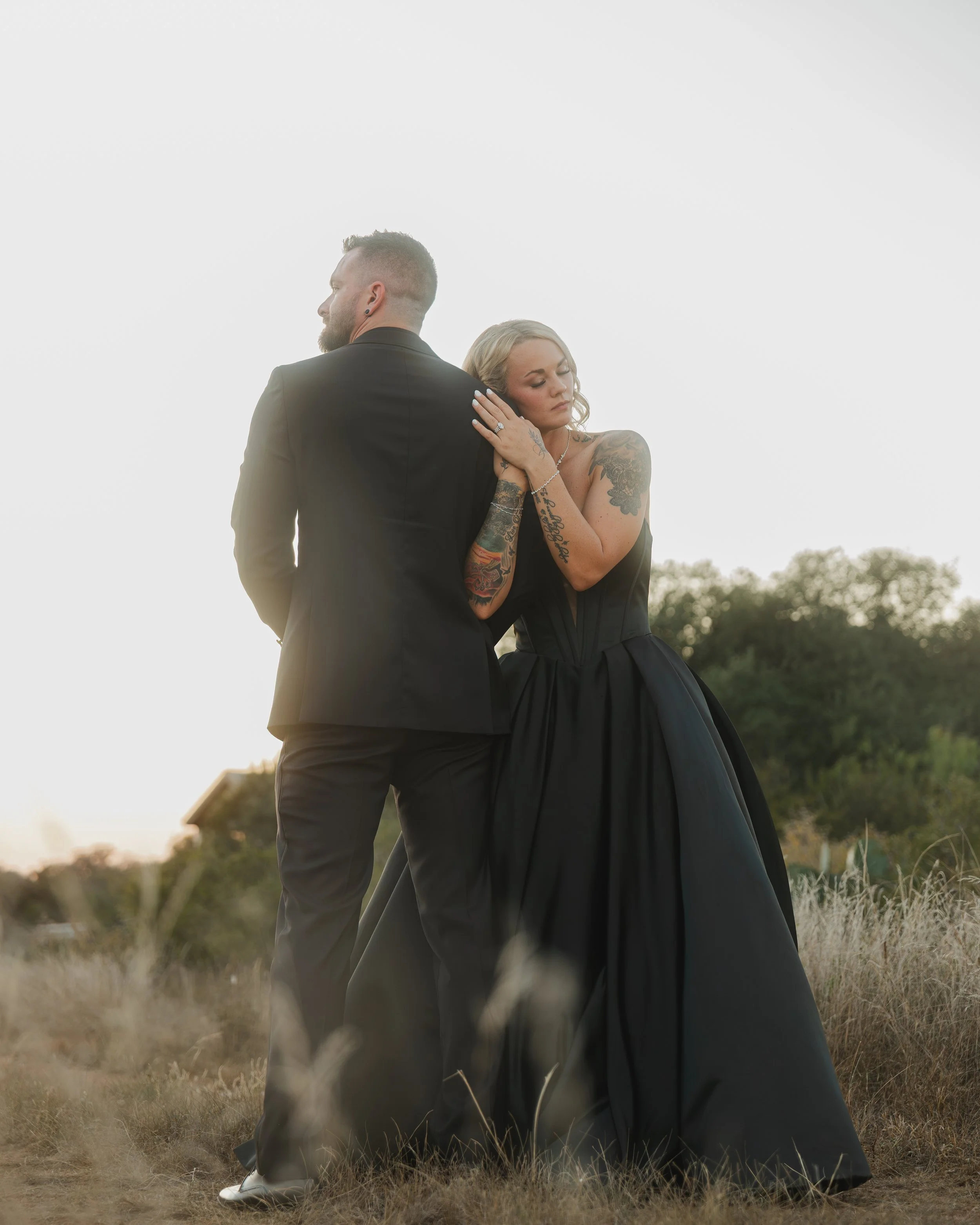 A man in a black suit and a woman in a black gown standing in a grassy field with trees in the distance, at sunset.