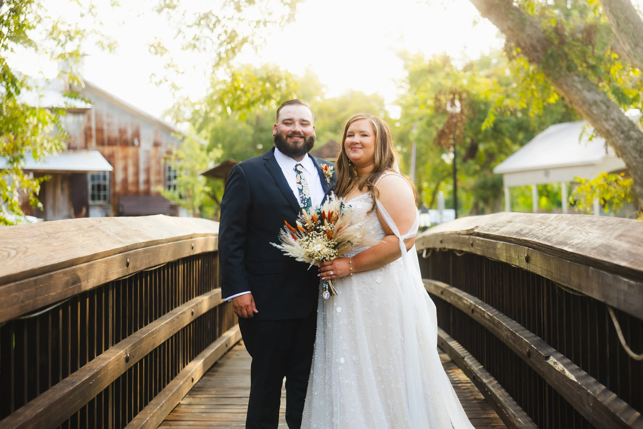 A smiling couple, a man in a dark suit with a floral tie and a woman in a white wedding dress holding a bouquet, standing on a wooden bridge outdoors during daytime.
