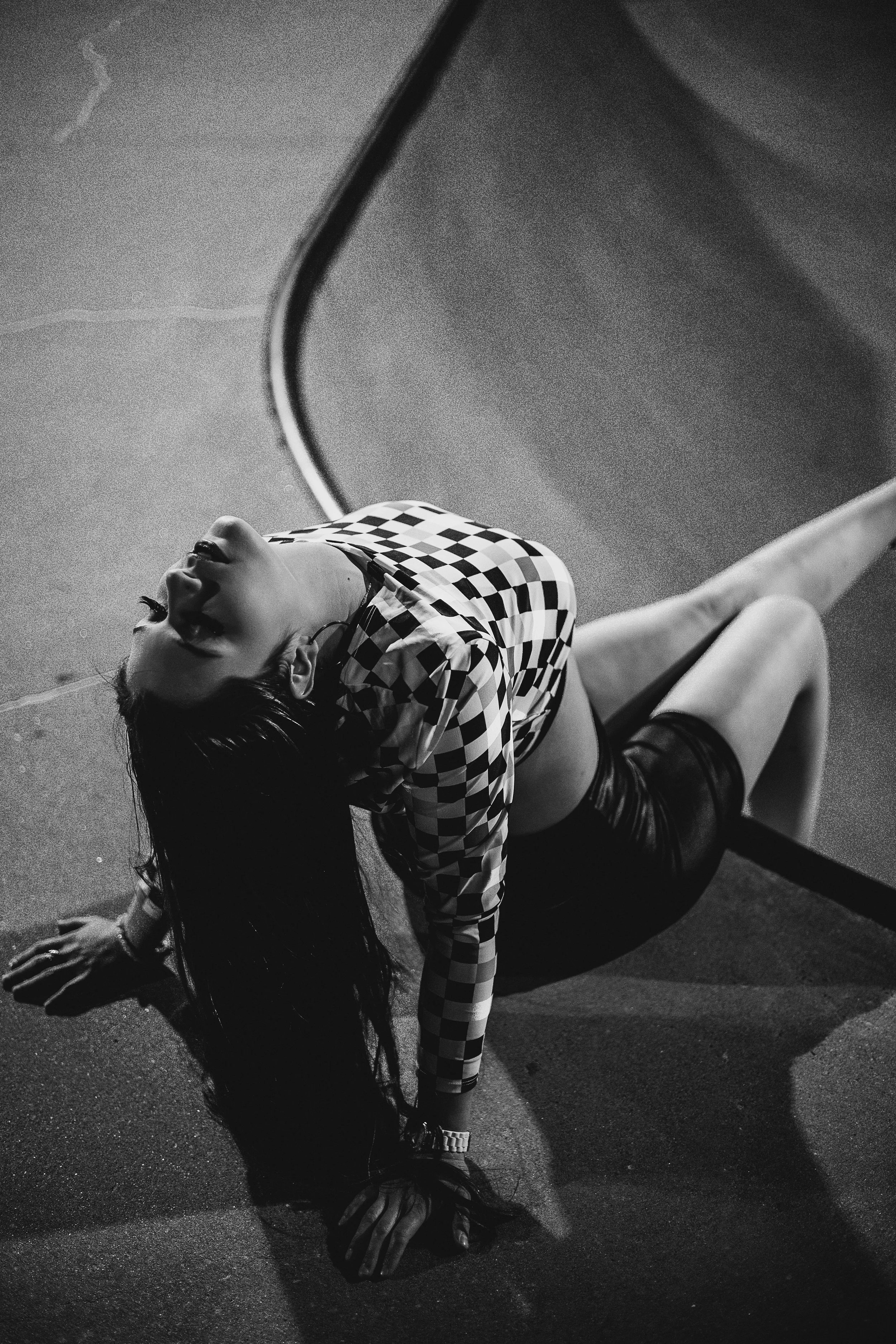 A woman with long dark hair, wearing a checkered shirt and shorts, reclines on the ground with her eyes closed and an expression of relaxation.