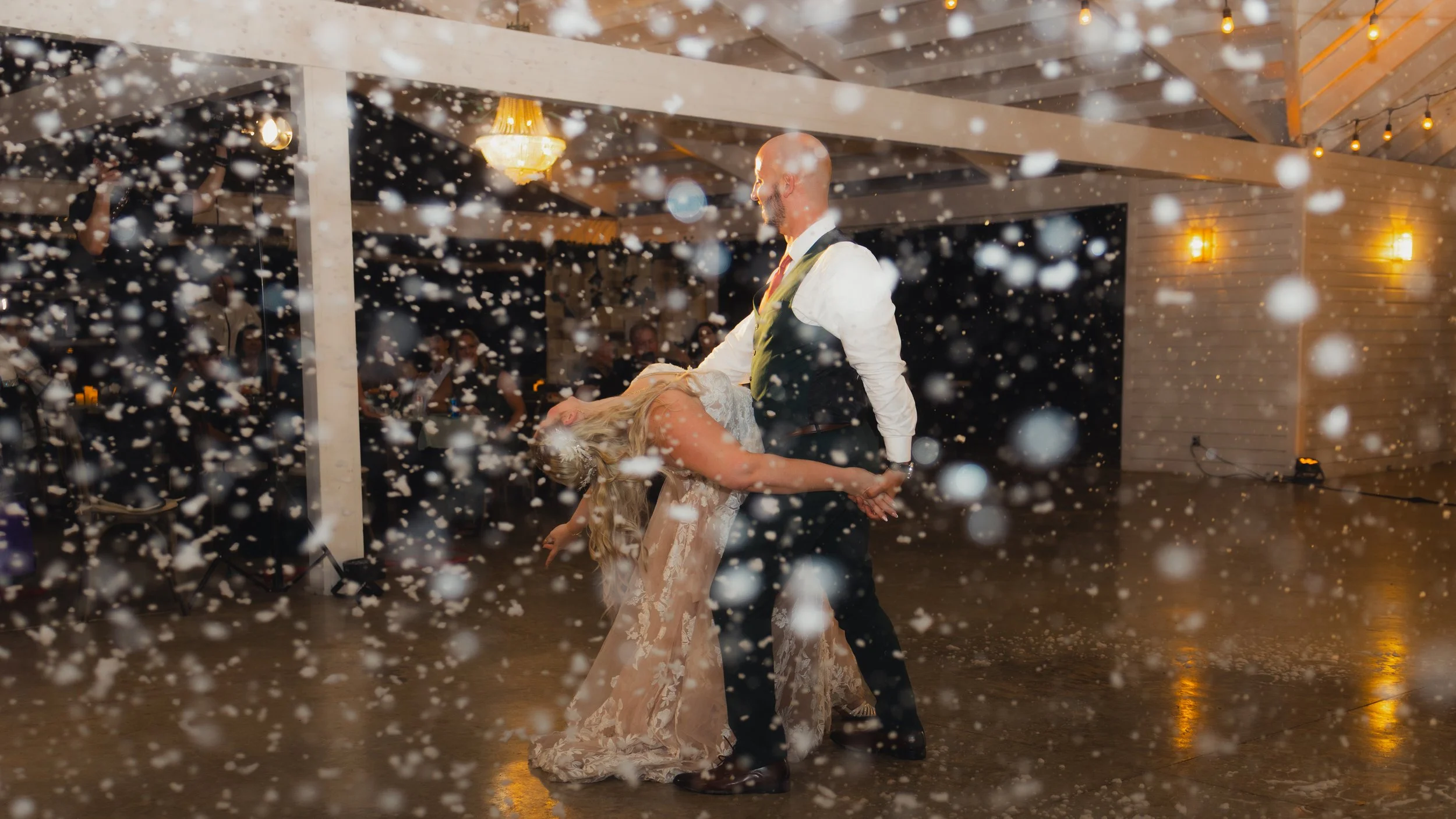 A couple dancing at their wedding reception with snow-like confetti falling around them. The bride is in a white lace gown, and the groom is in a dark suit with a white shirt. They are holding hands and smiling, while guests watch in the background u