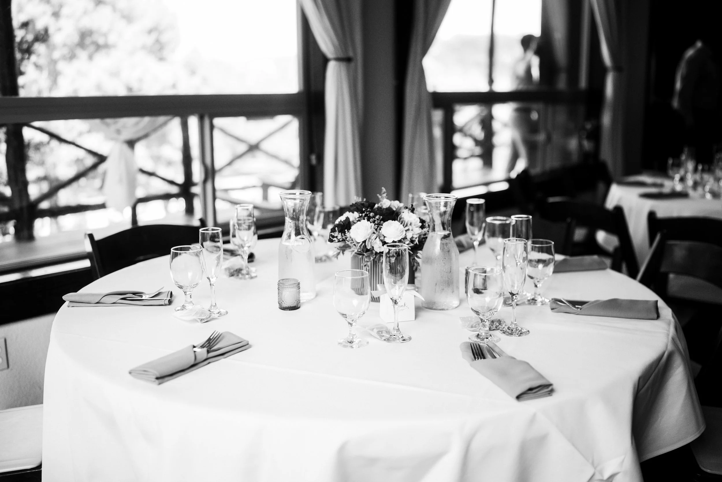 Round table set with wine glasses, water glasses, folded napkins, a flower centerpiece, and glass bottles in a room with large windows and curtains.