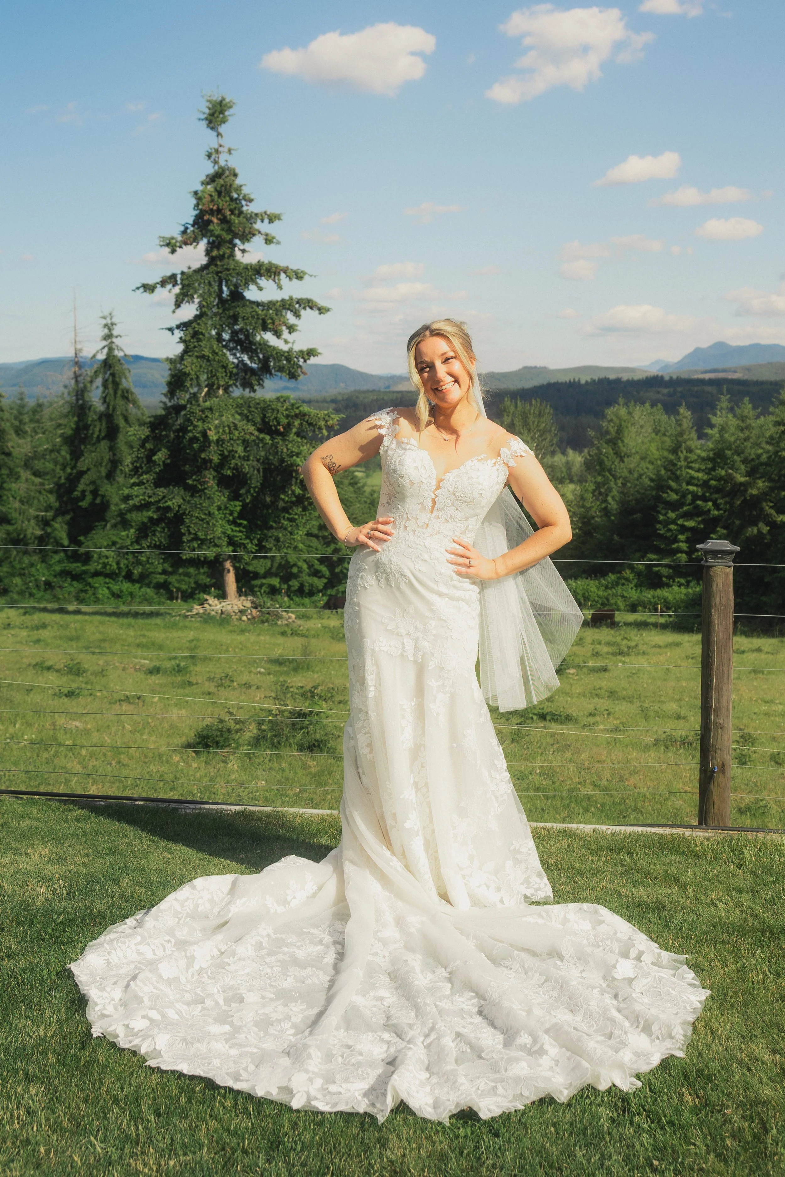 A smiling woman in a white wedding dress standing on a grassy field with mountains and trees in the background under a partly cloudy sky.