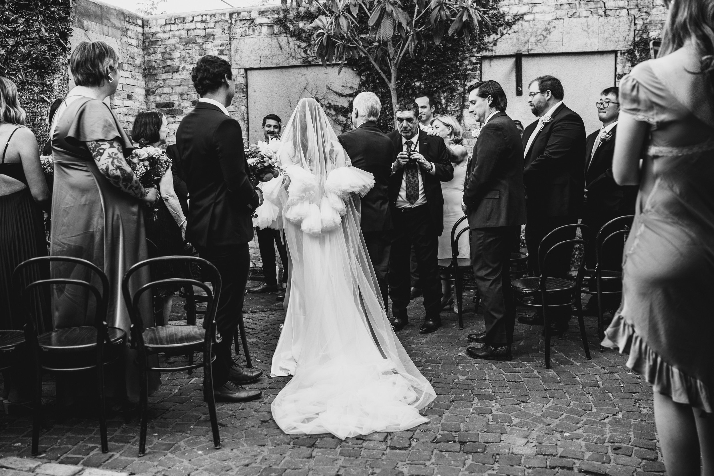 A black-and-white photo of a wedding ceremony in a courtyard, with guests standing around a bride and groom who are facing each other. The bride is wearing a long white dress with a veil, and the groom is in a dark suit. The guests are dressed in for