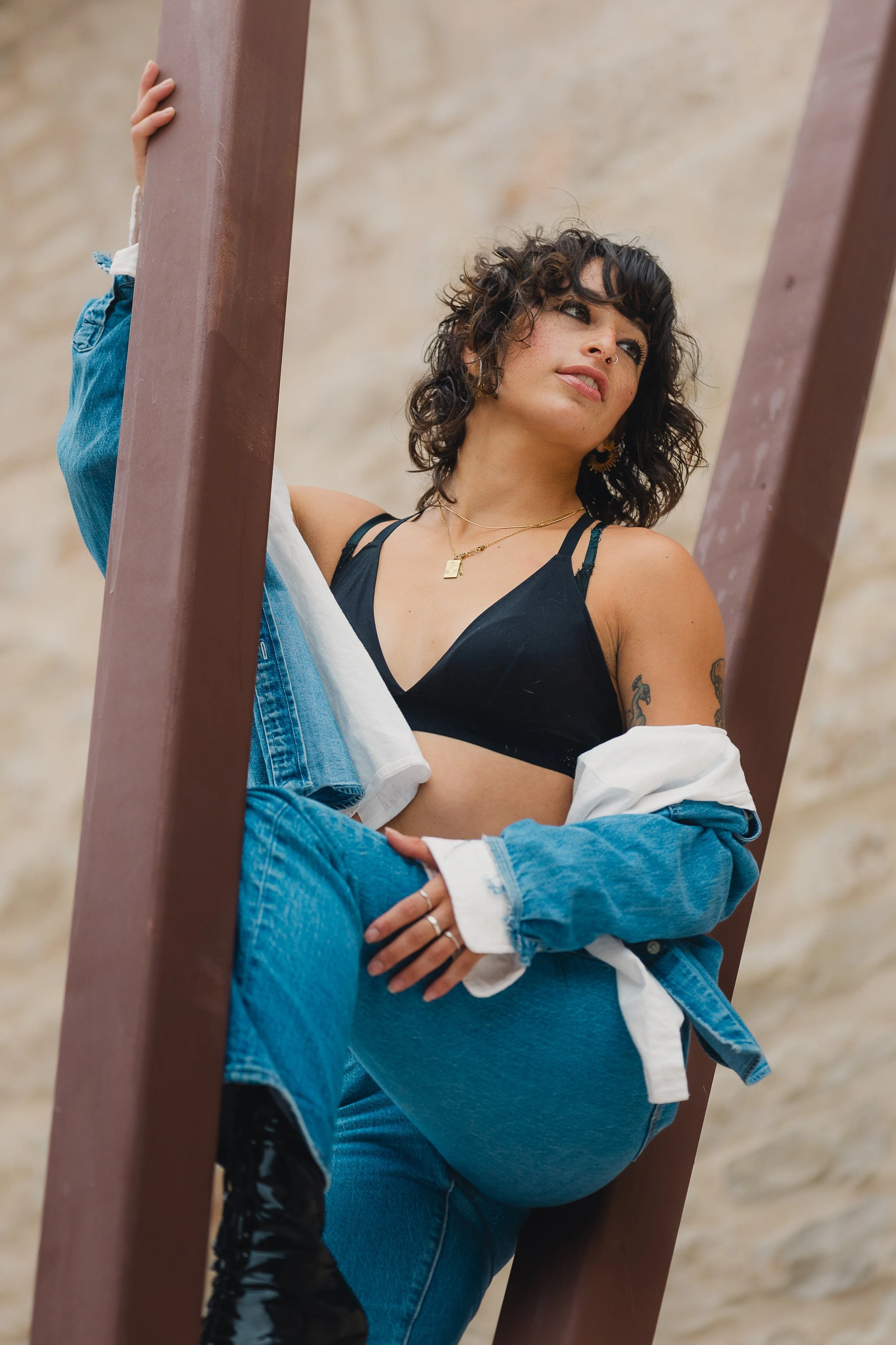 A young woman with dark, curly hair, wearing a black bralette, blue jeans, and a denim jacket, poses against a metal structure outdoors.
