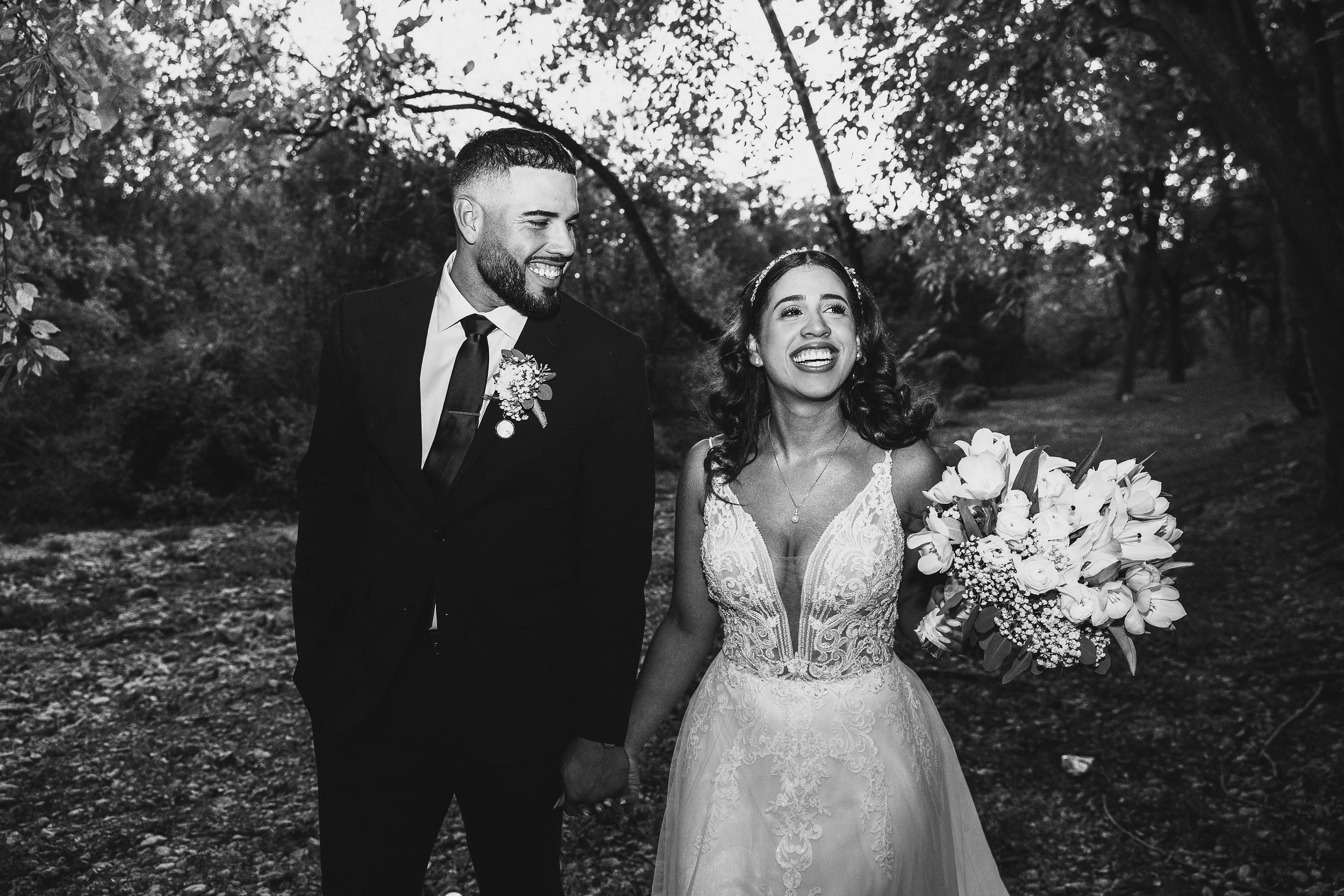 Black and white photo of a smiling couple holding hands in a wooded outdoor setting, with the woman holding a large bouquet of flowers.