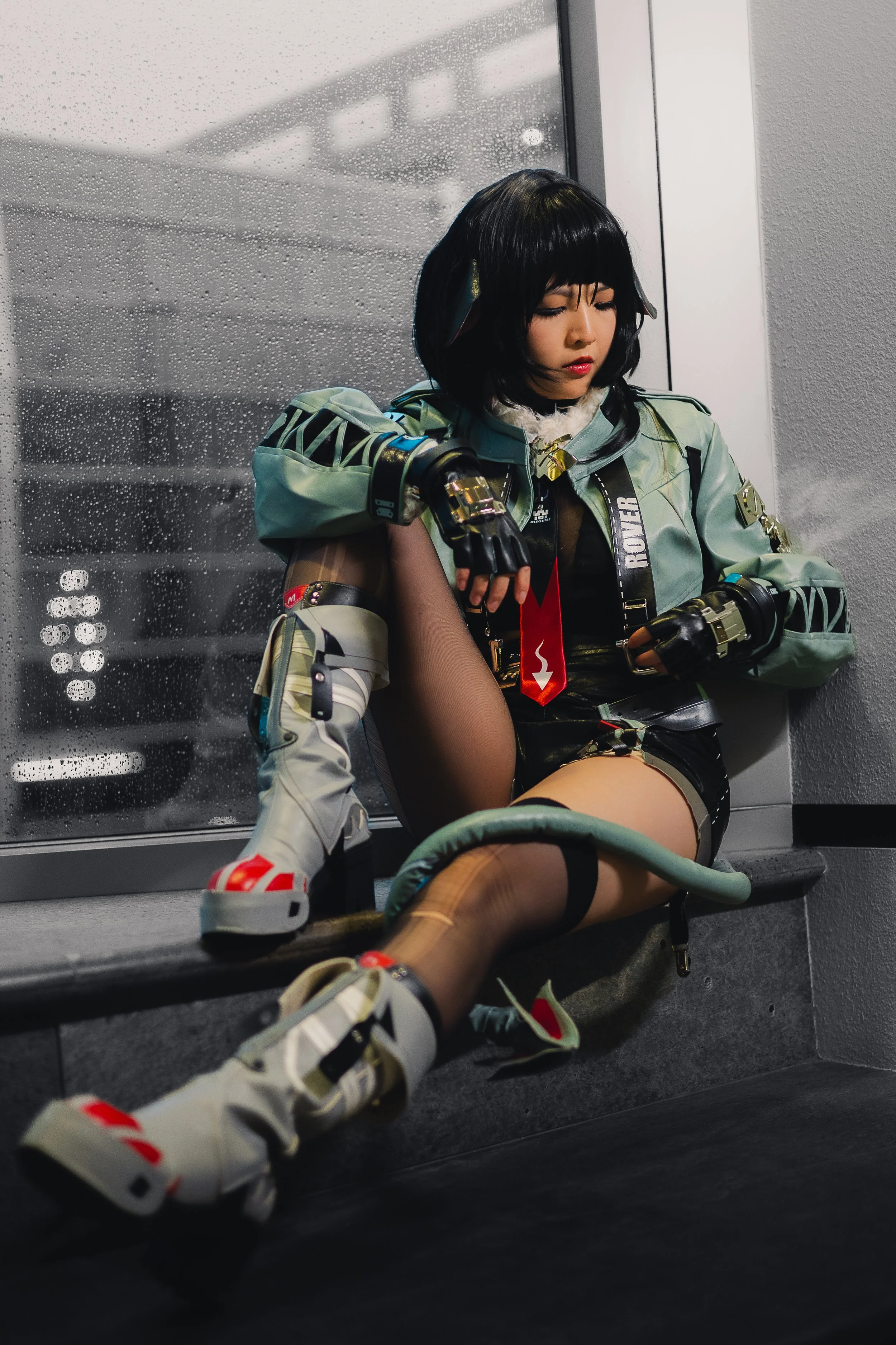A woman sits on a bench in front of a rain-streaked window, wearing a futuristic military-style jacket, shorts, and high-tech footwear, with a serious expression.