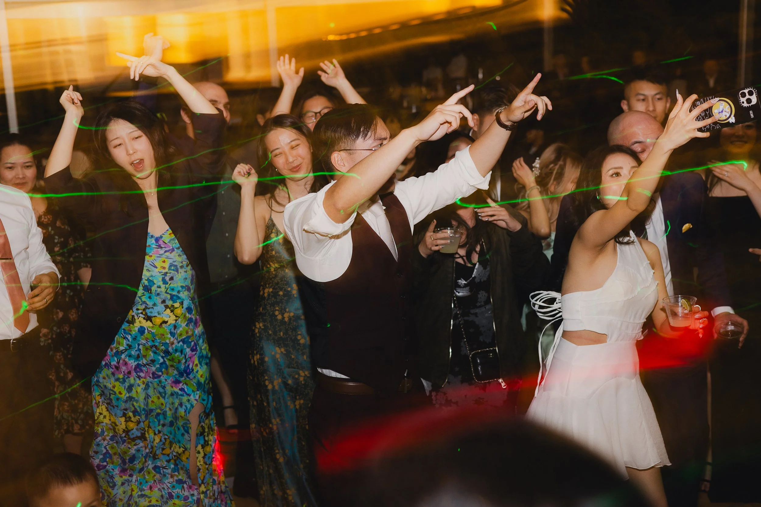A group of people dancing at a party or celebration, with some holding drinks, colorful lighting effects, and lively expressions.