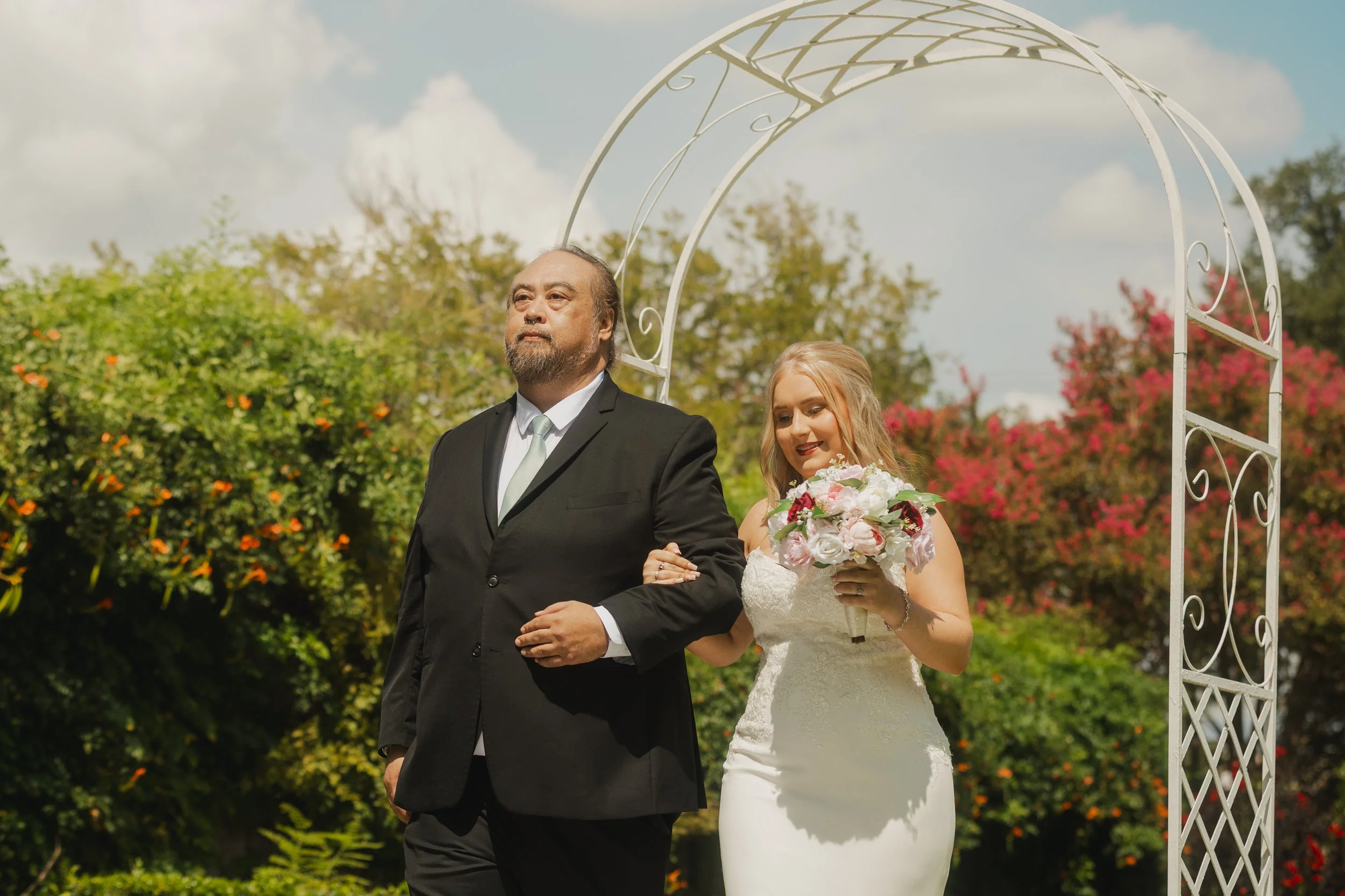 A bride with blonde hair in a white wedding dress holding a bouquet of pink and white roses walking arm-in-arm with a man in a black suit at an outdoor wedding ceremony under a white decorative arch, with colorful trees and a partly cloudy sky in the