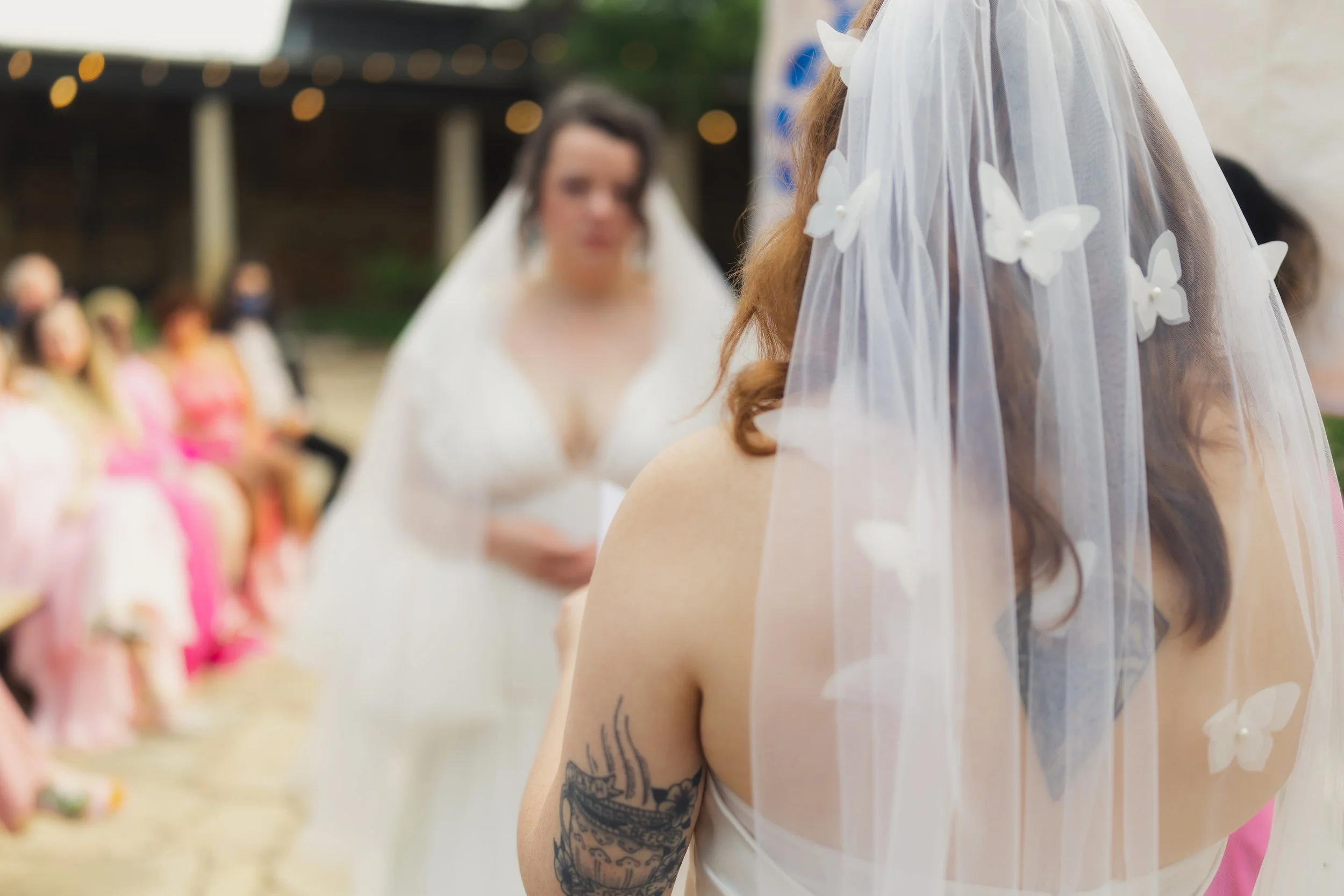 Close-up of a woman with long brown hair and a tattoo on her arm, wearing a white veil with butterfly embellishments at a wedding ceremony. In the background, another woman in a white dress, likely the bride, and several guests seated, some wearing m