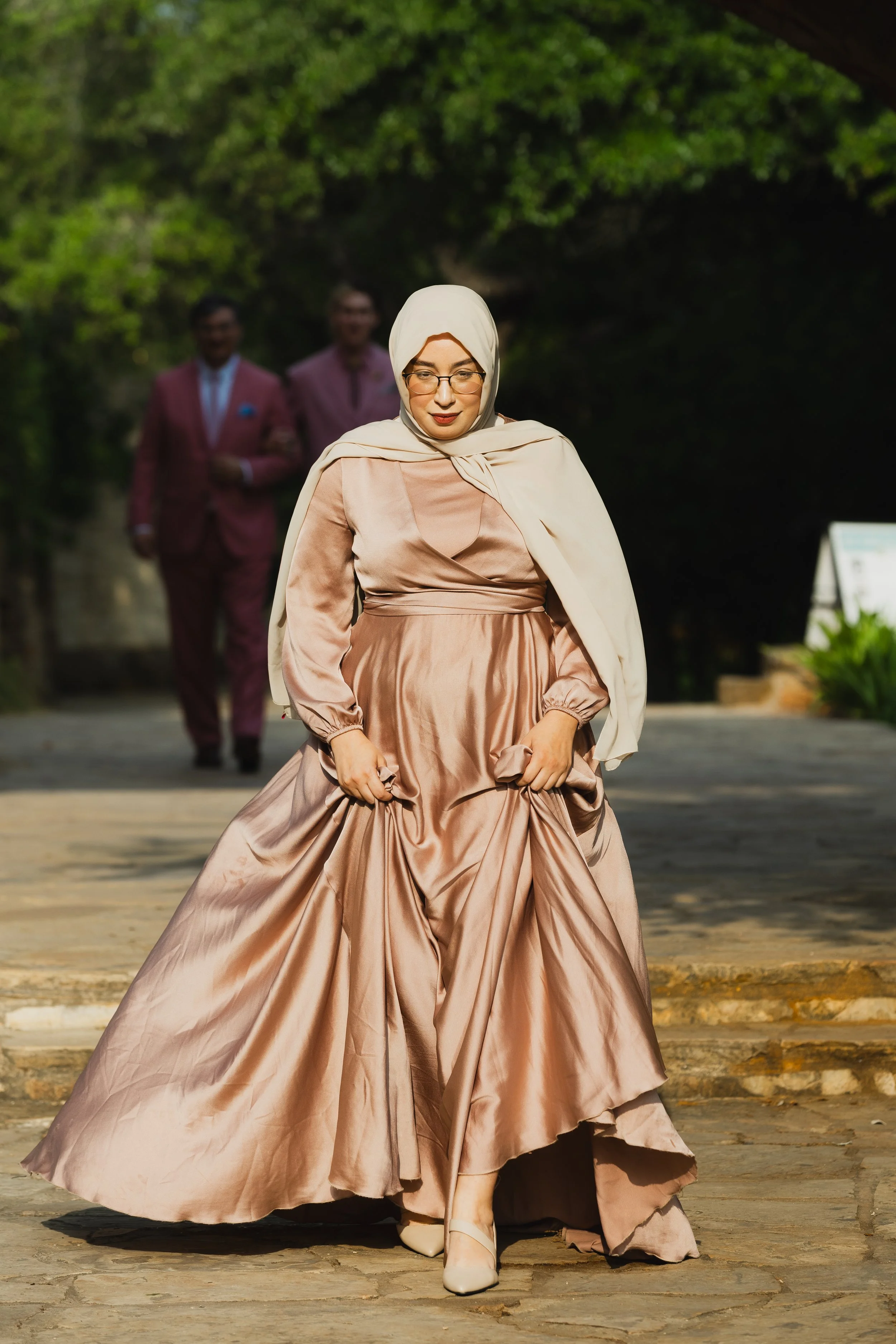 Woman wearing a beige hijab and a long, flowing satin gown walking outdoors with two men in suits visible in the background.