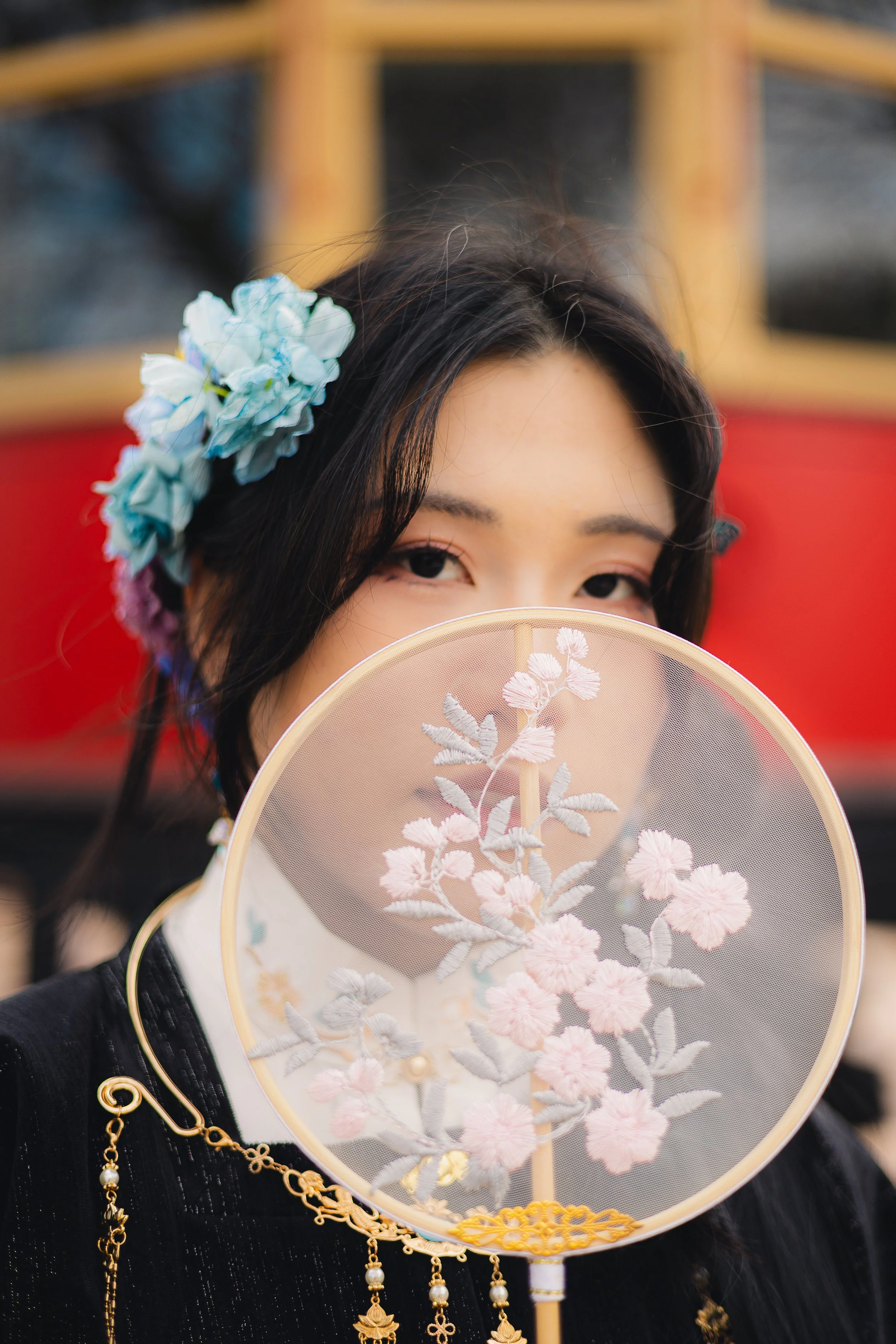 A woman holding a decorative fan with pink floral embroidery partially covering her face, wearing a black outfit with gold jewelry, and a blue flower in her dark hair.