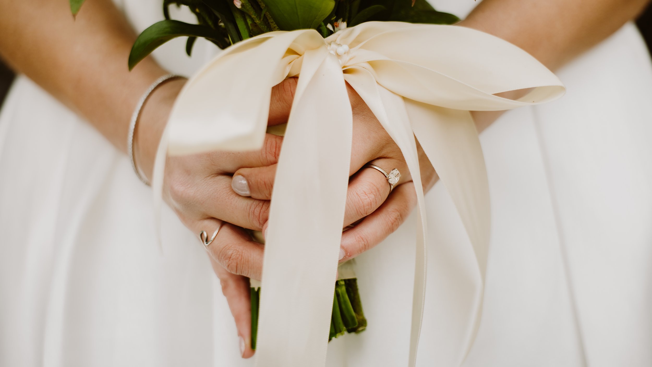 A person holding a bouquet of white flowers with ribbons, wearing a silver ring and a simple bracelet.