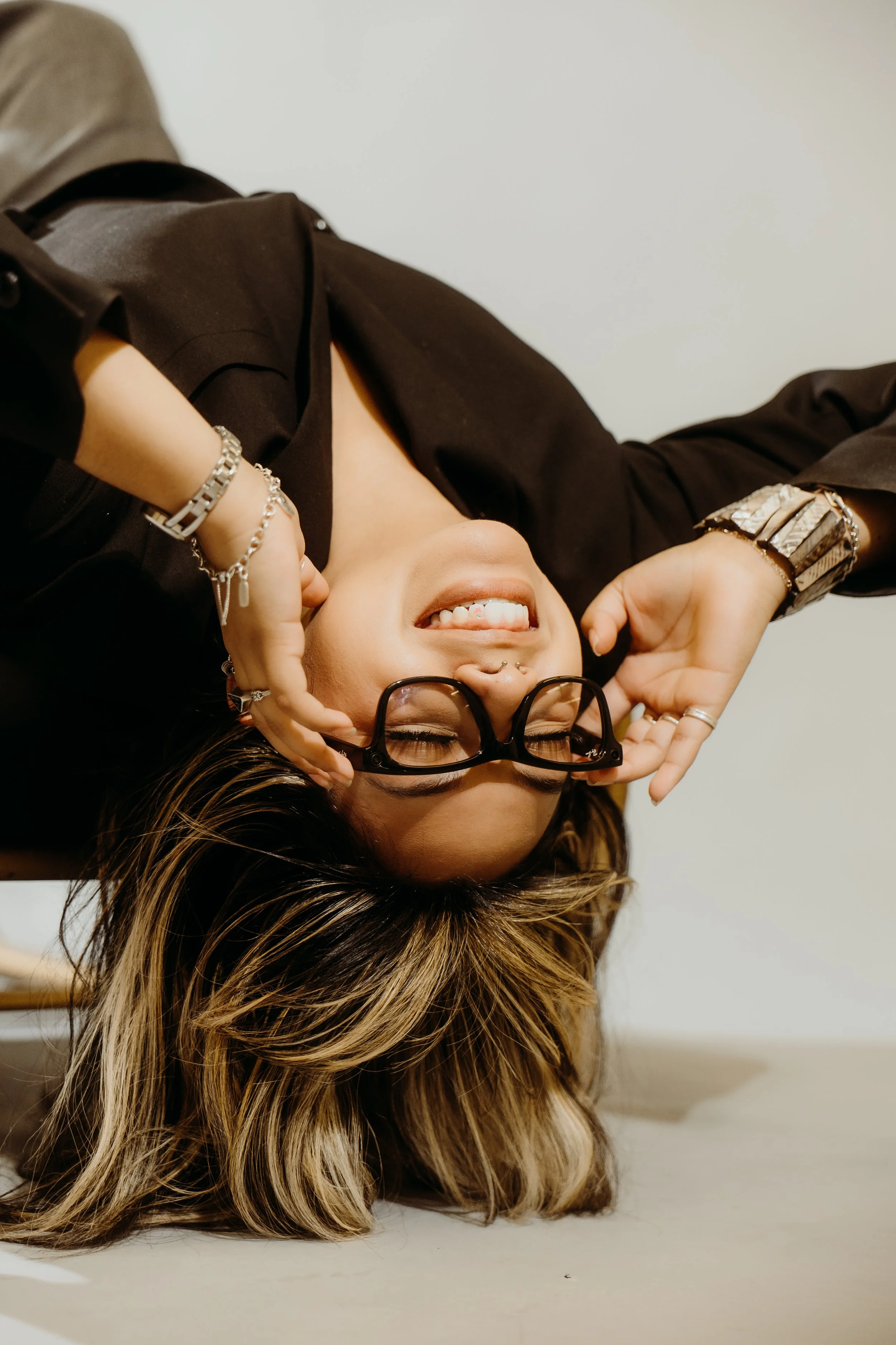 A woman lying on her back with her hair spread out, smiling, with glasses held upside down over her eyes, wearing jewelry, a black shirt, and a septum piercing.