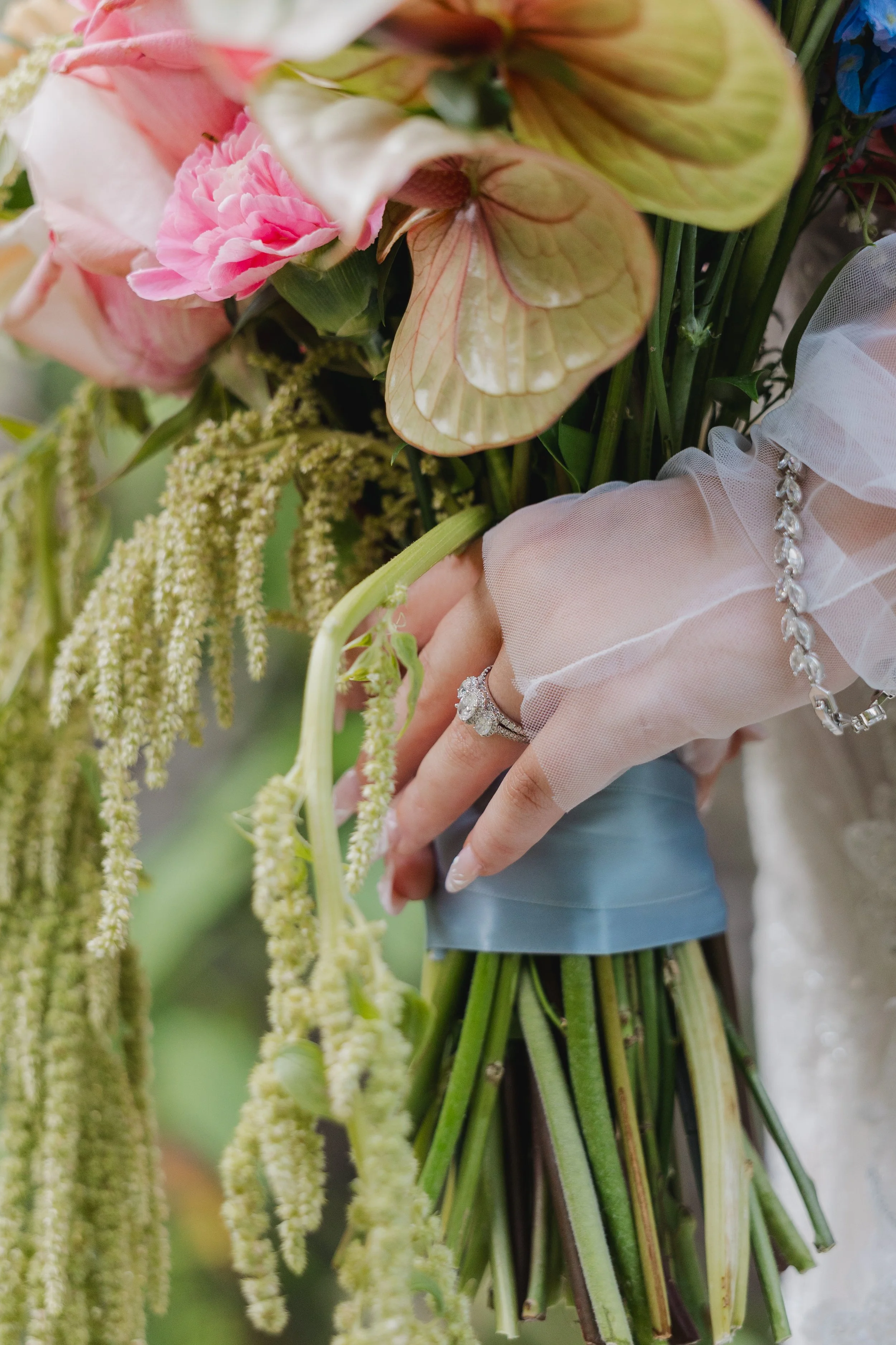 Close-up of a bride holding a bouquet of pink and cream flowers with green foliage, wearing a diamond engagement ring and a sheer, ruffled sleeve.
