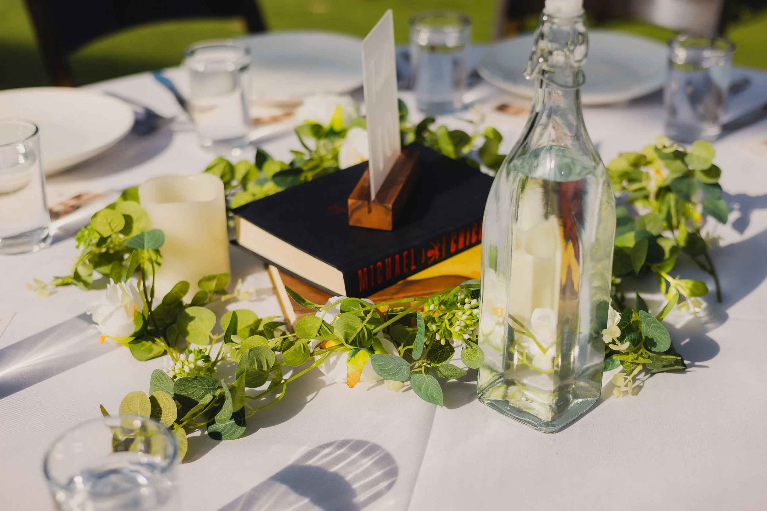 Table centerpiece with a black book, a white candle, a glass bottle with a white candle inside, surrounded by green foliage and white flowers, set on a white tablecloth at an outdoor event.
