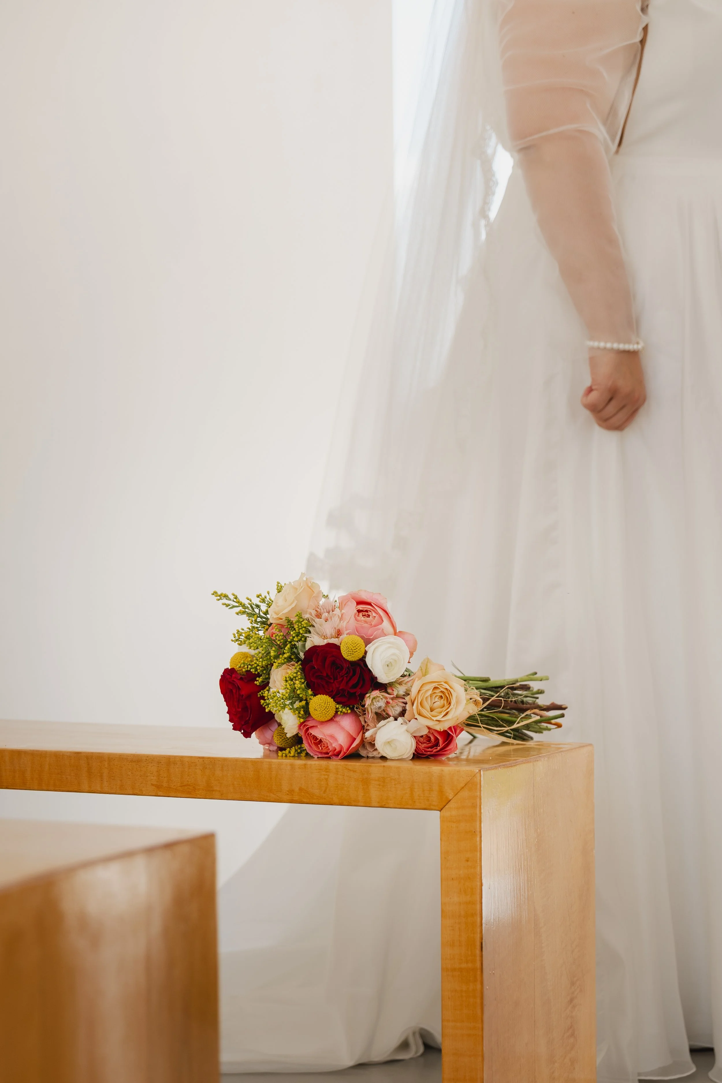 Close-up of a wedding bouquet with pink, red, white, and yellow flowers resting on a wooden table, with a bride in a white wedding dress and pearl bracelet in the background.