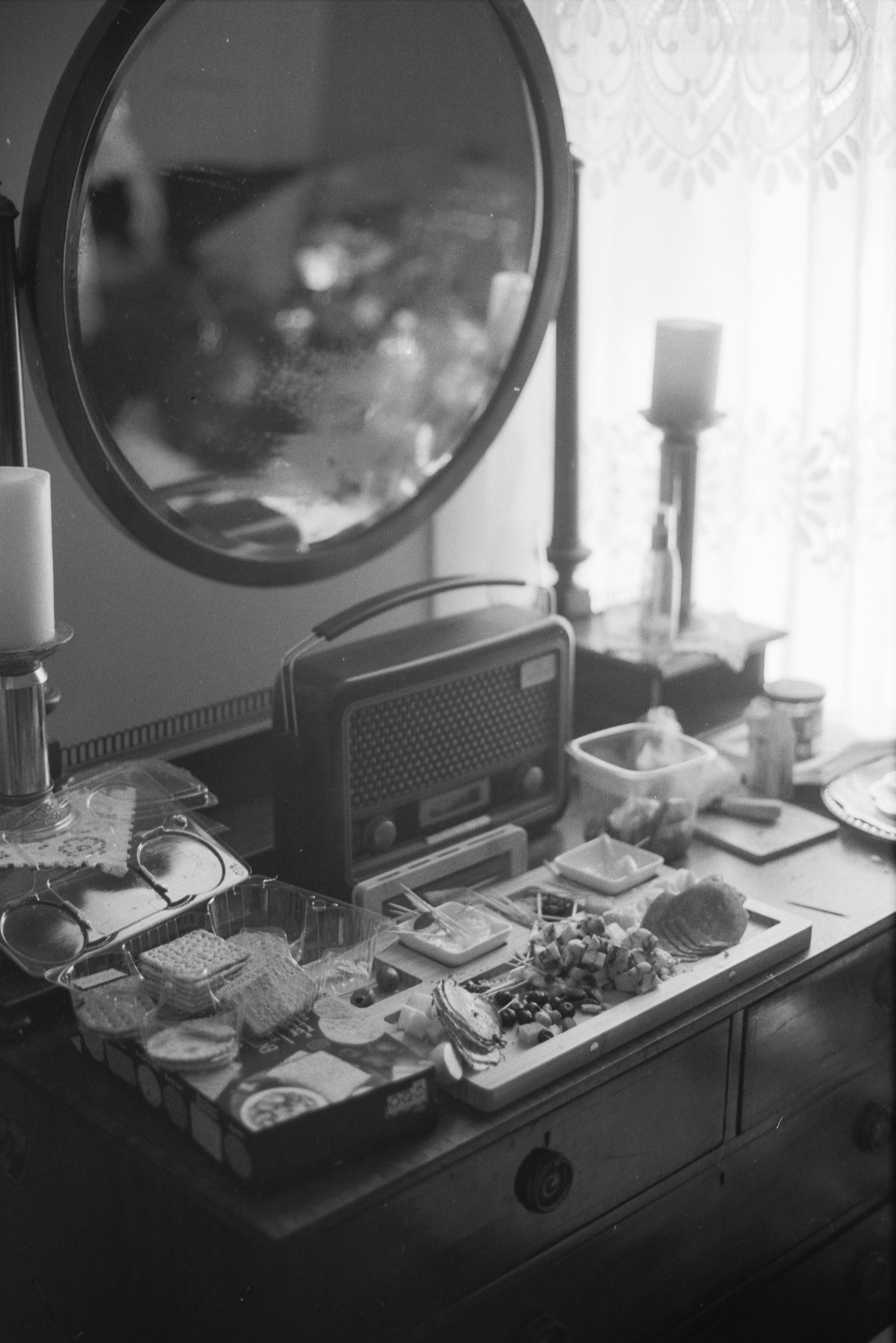 A cluttered dresser with a mirror, candles, a radio, and various food items including crackers, cheese, and snacks.