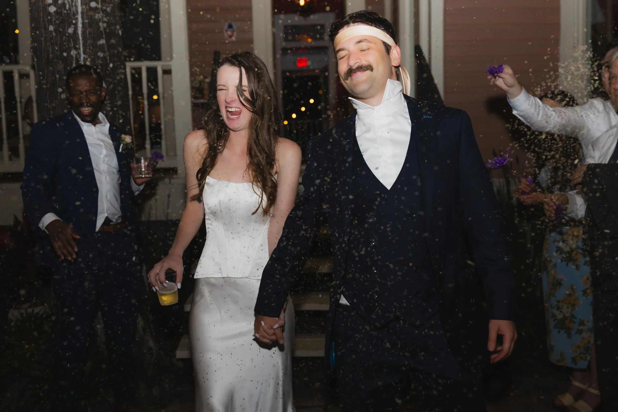 Couple holding hands and celebrating at their wedding reception with friends, confetti falling, and joyful expressions.