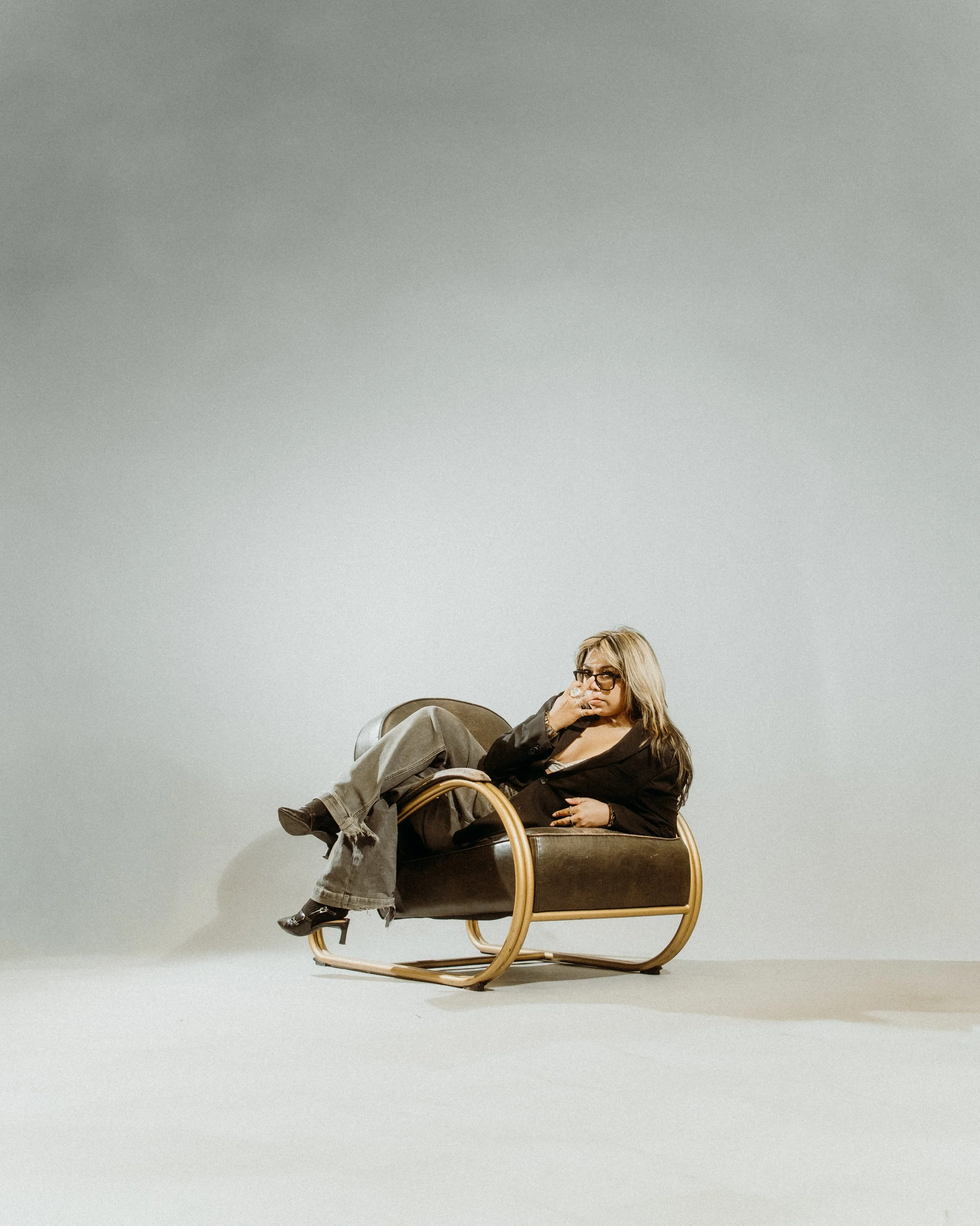 Woman with blond hair, glasses, dark clothing, sitting on a modern brown and gold chair, posing against a plain white background.