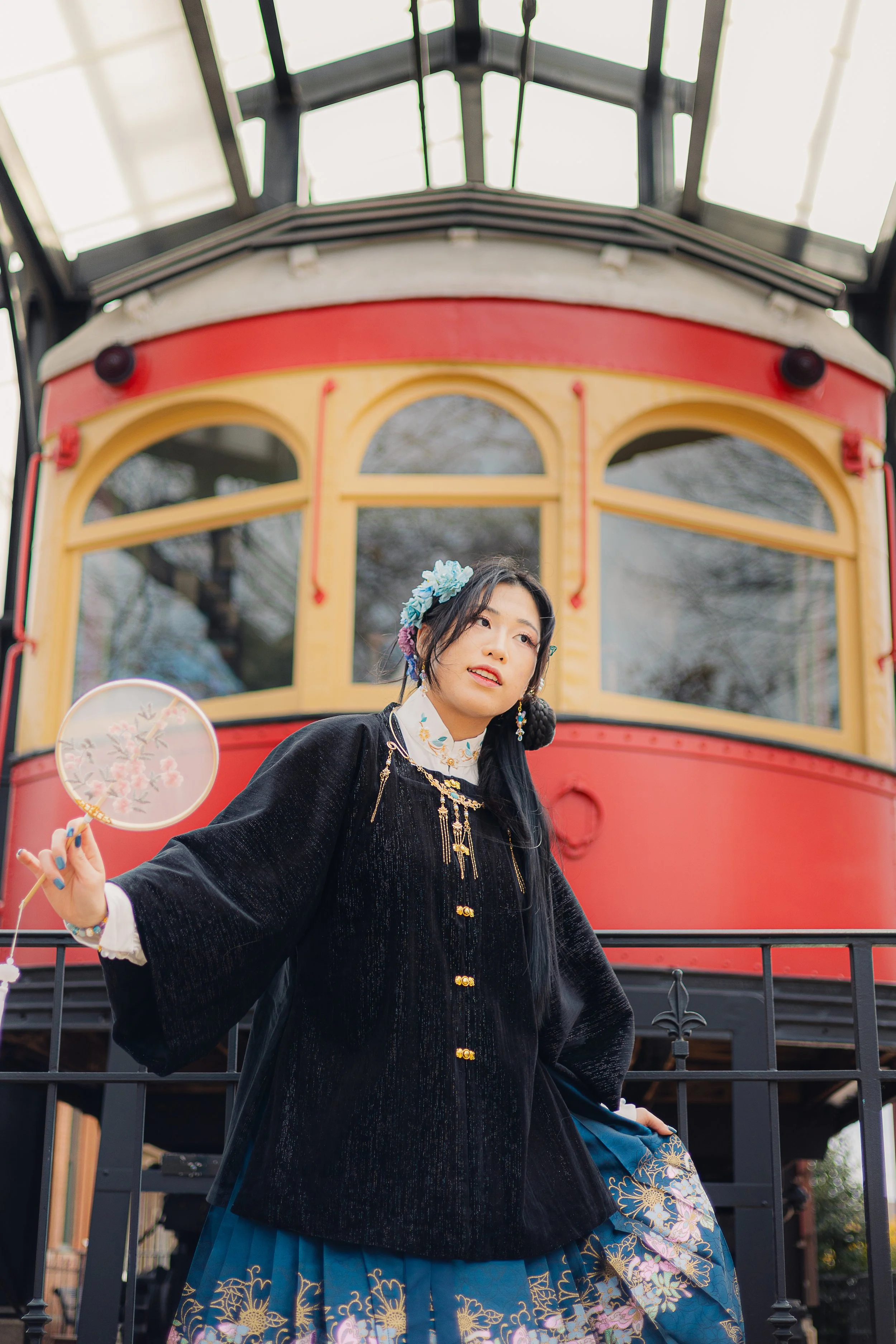 A woman dressed in traditional Asian attire standing in front of a vintage red and yellow tram.