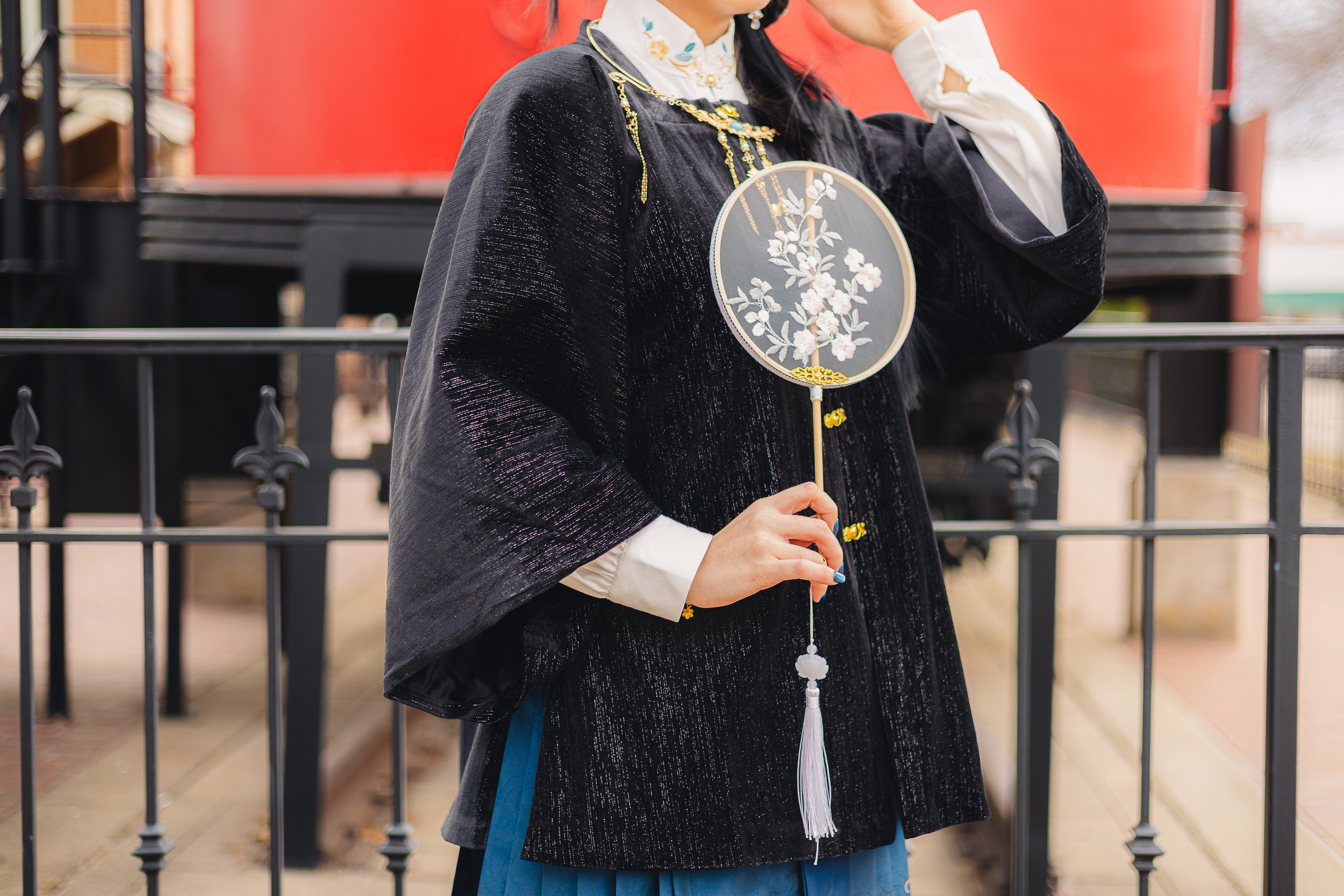 Person holding a decorative circular fan with floral embroidery, dressed in traditional or cultural attire, standing outdoors behind a black metal fence with a red structure in the background.