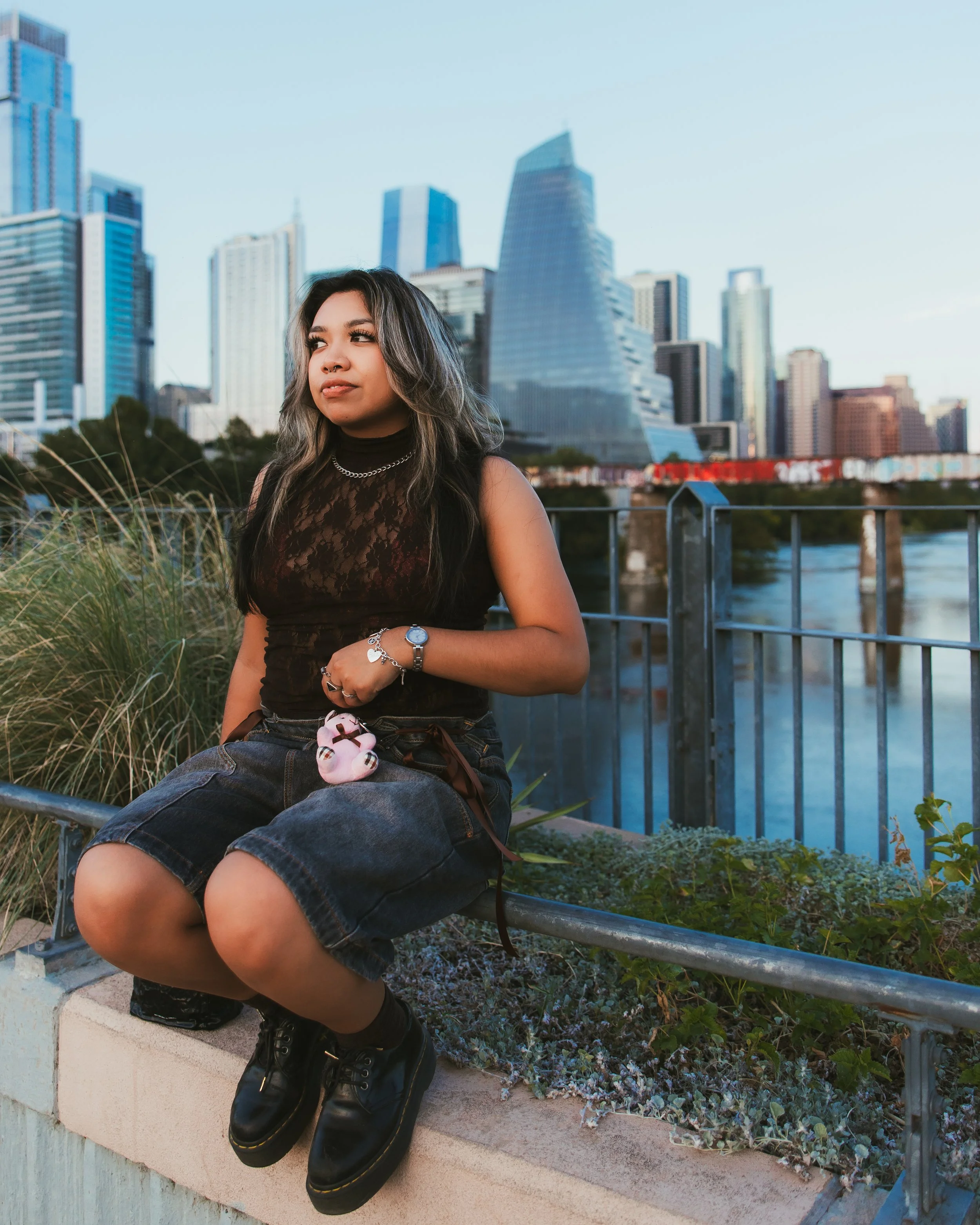 Young woman sitting on a ledge near a river with a city skyline in the background during daytime.