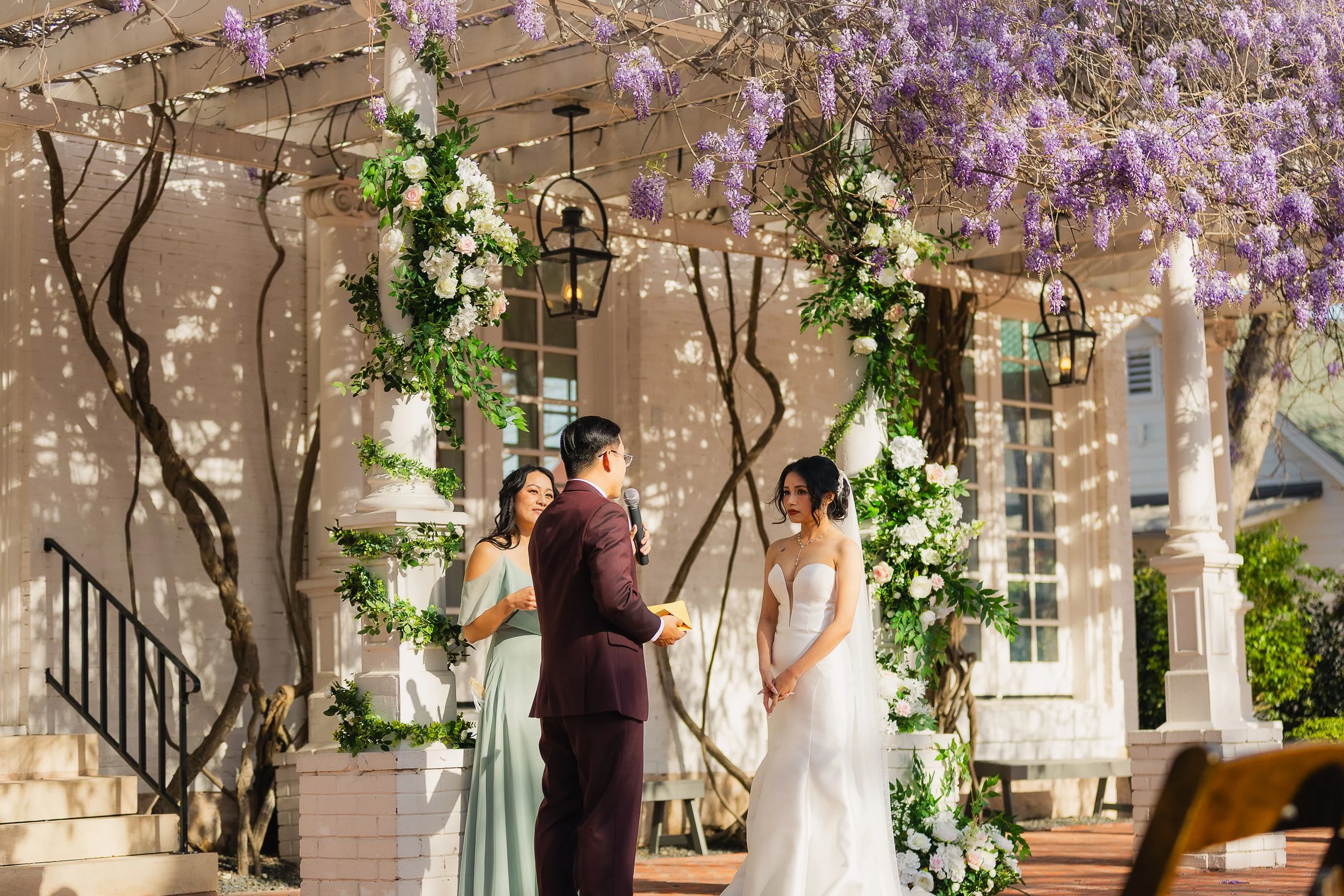 A wedding ceremony outdoors with a bride in a white dress and a groom in a maroon suit under a floral arch decorated with purple and white flowers. An officiant and an officiant assisting are present. The setting includes a gazebo with hanging lanter