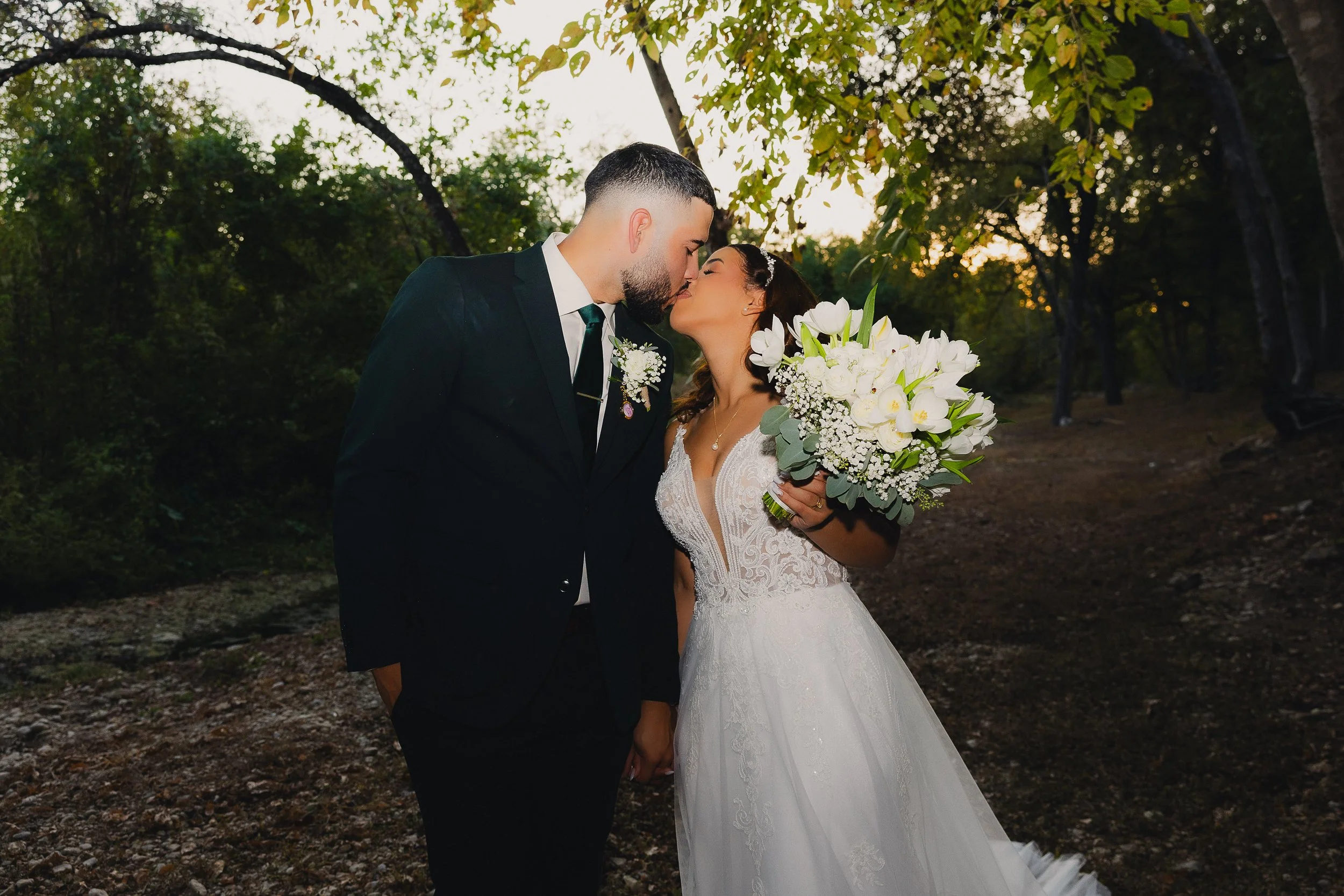 A bride and groom kiss outdoors during their wedding, with the bride holding a large bouquet of white flowers and greenery, and trees in the background at sunset.