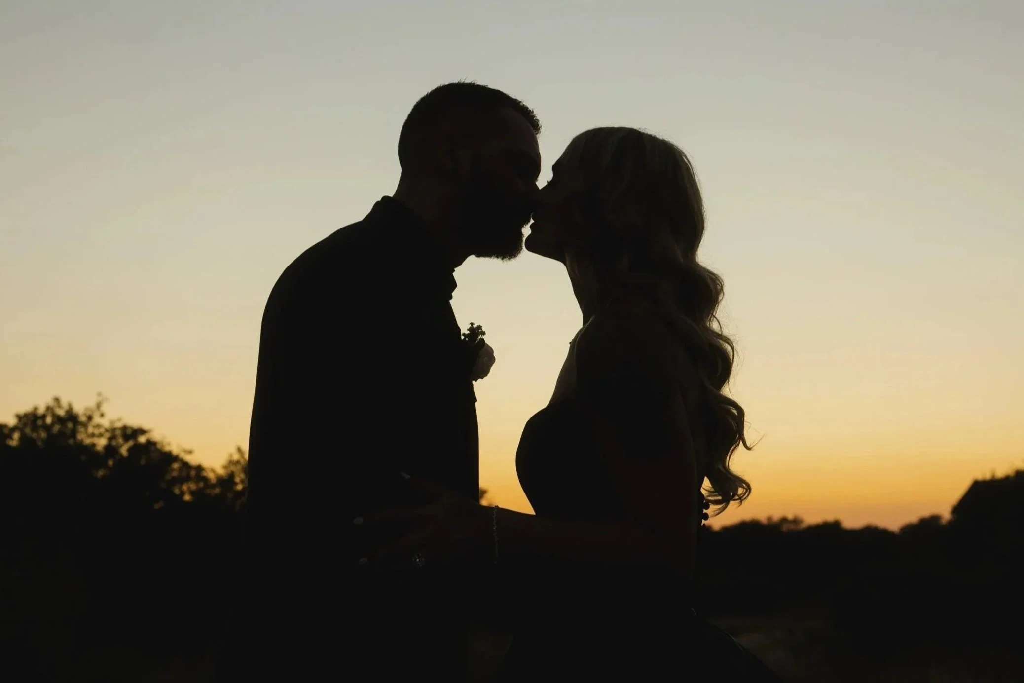 Silhouette of a couple, man and woman, sharing a kiss during sunset in an outdoor setting.