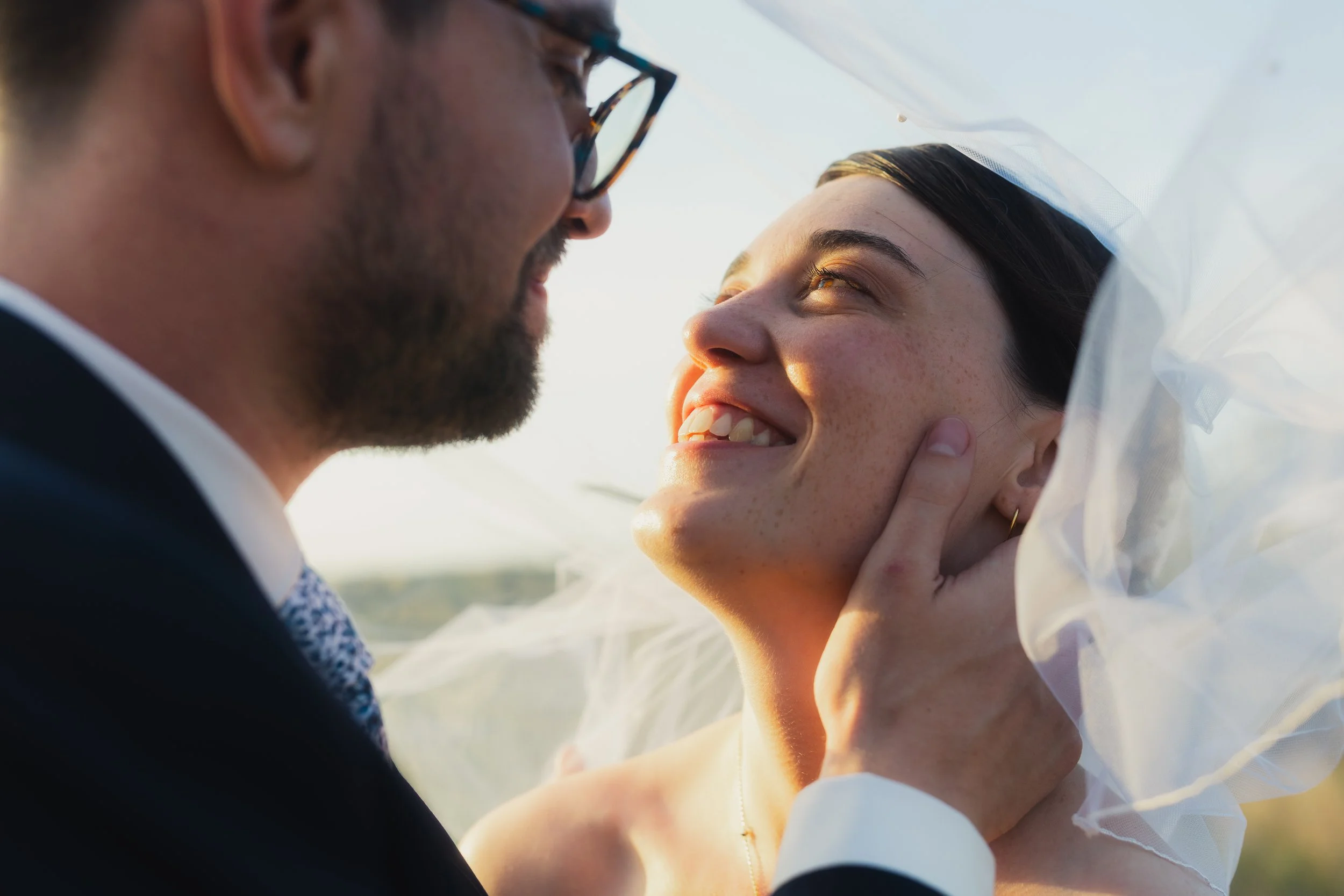 A close-up of a couple, with the man gently holding the woman's face, smiling at each other under a veil in a wedding setting during sunset.