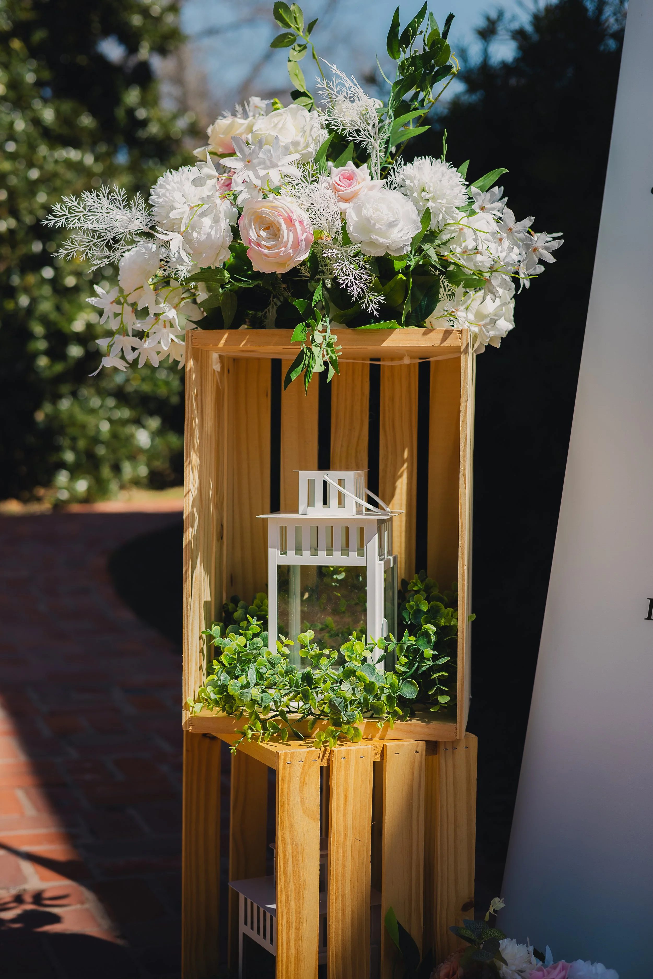 Flower arrangement with white and pink roses, white lilies, and greenery in a wooden box, with a small white decorative lantern and green plants underneath, set outdoors on a brick path.