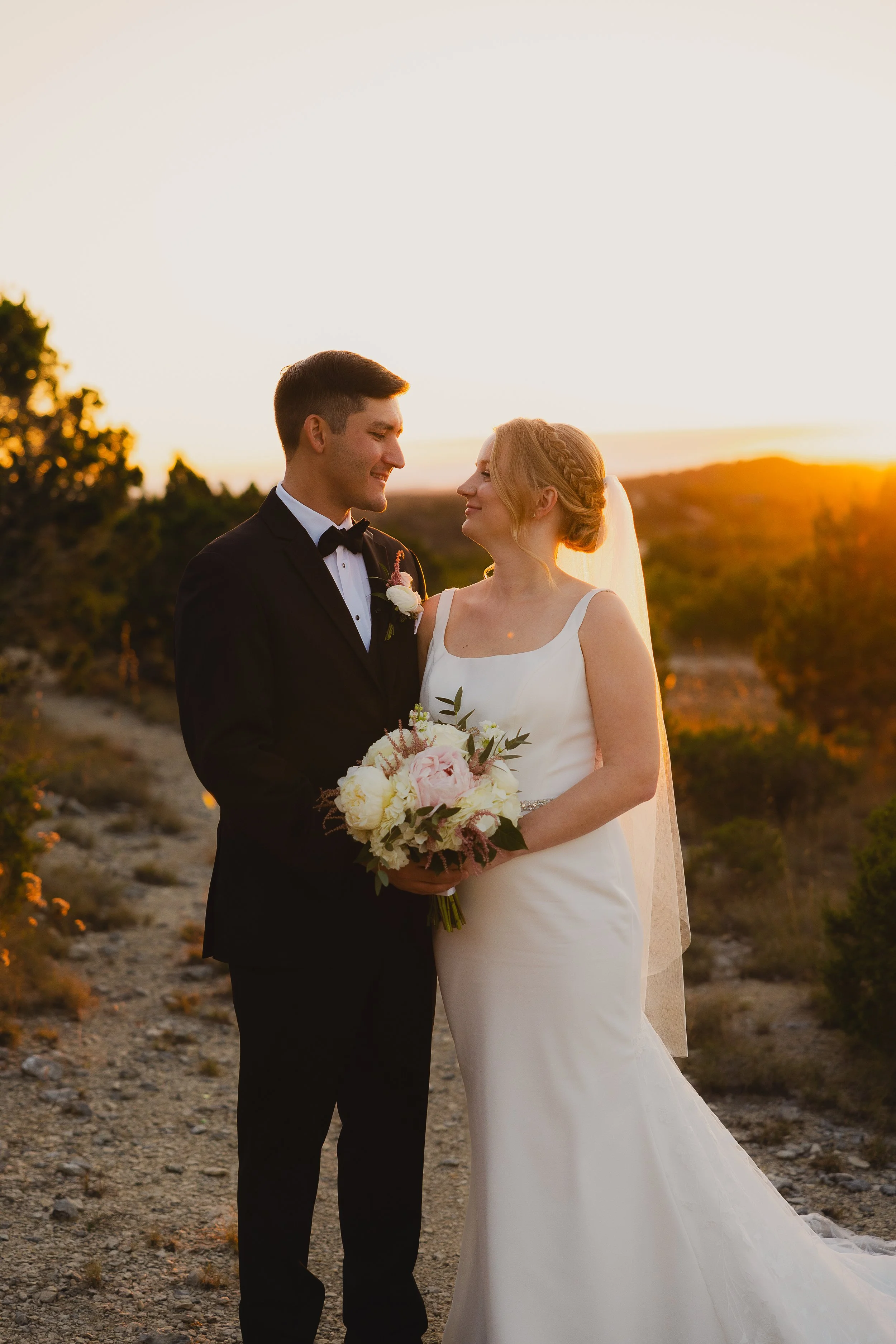A bride and groom standing together outdoors at sunset, smiling at each other. The bride is holding a bouquet of flowers, wearing a white wedding gown and veil. The groom is dressed in a black tuxedo with a bow tie.