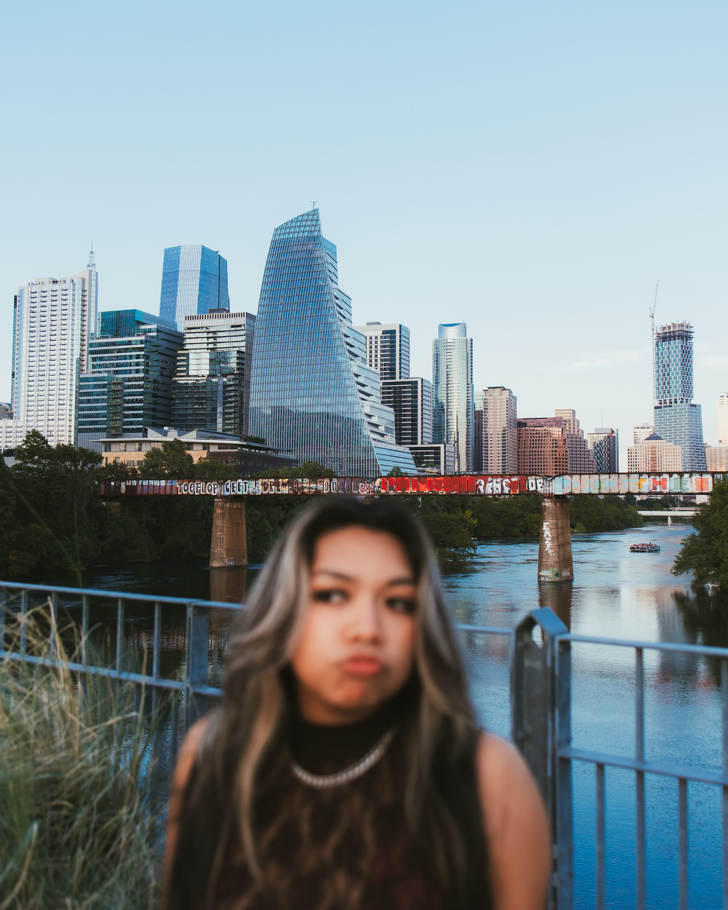 Blurred woman with long hair and a necklace in front of cityscape with tall buildings and a river.