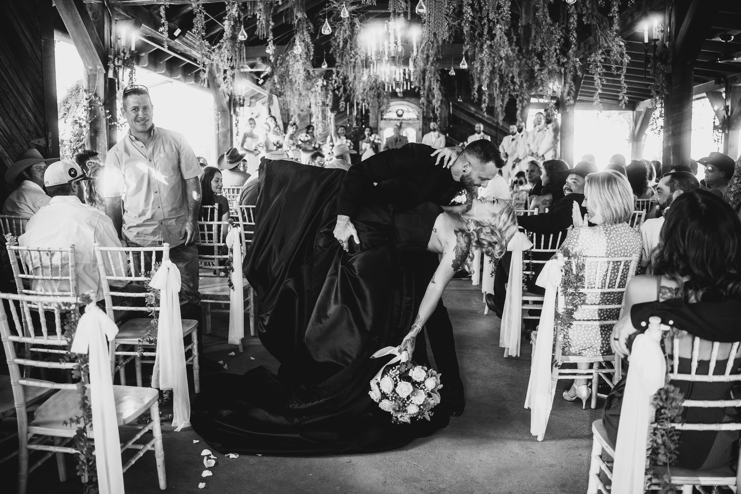 A black and white photo of a wedding ceremony where the bride and groom are sharing a kiss. The groom is dressed in a suit, and the bride is in a gown holding a bouquet of flowers. Guests are seated on either side watching the couple, with some smili