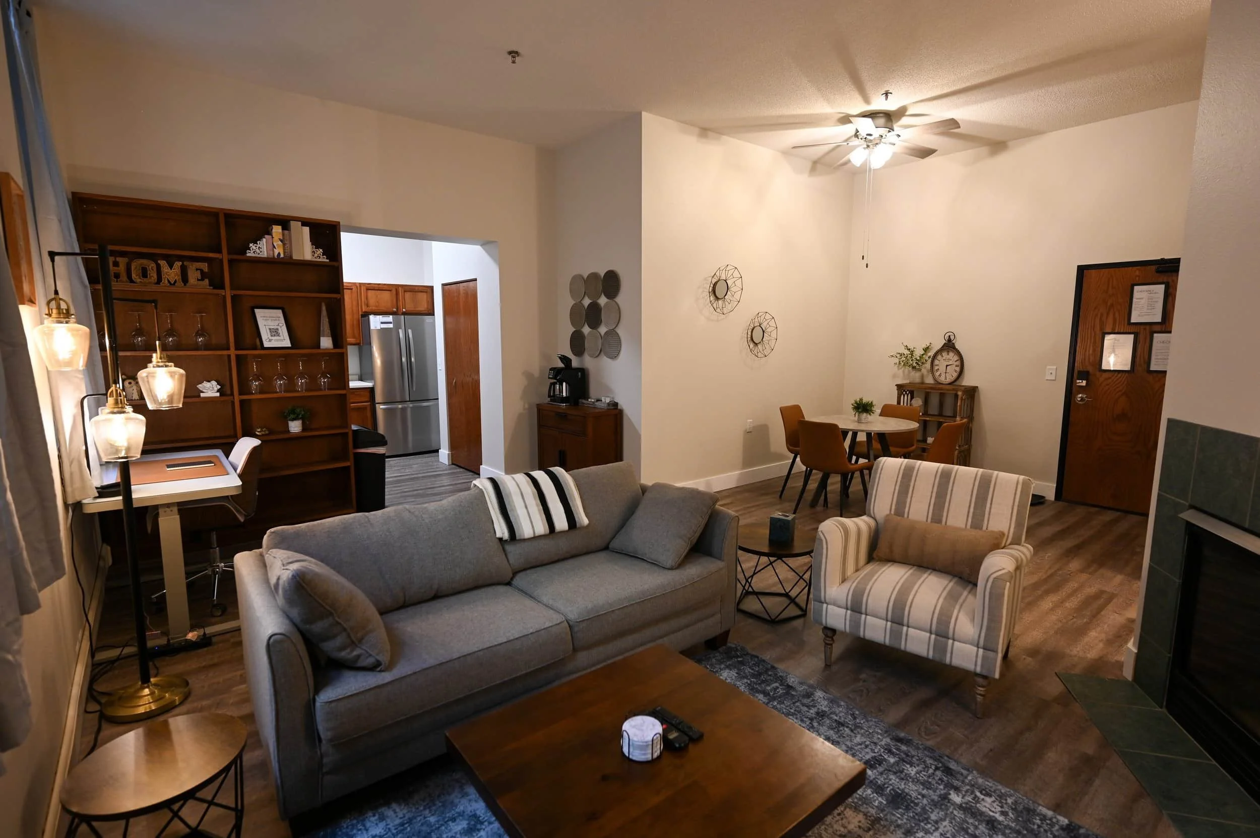 A cozy living room in apartments in Rochester with a gray sofa, striped armchair, dining area, kitchen, and desk with shelves.