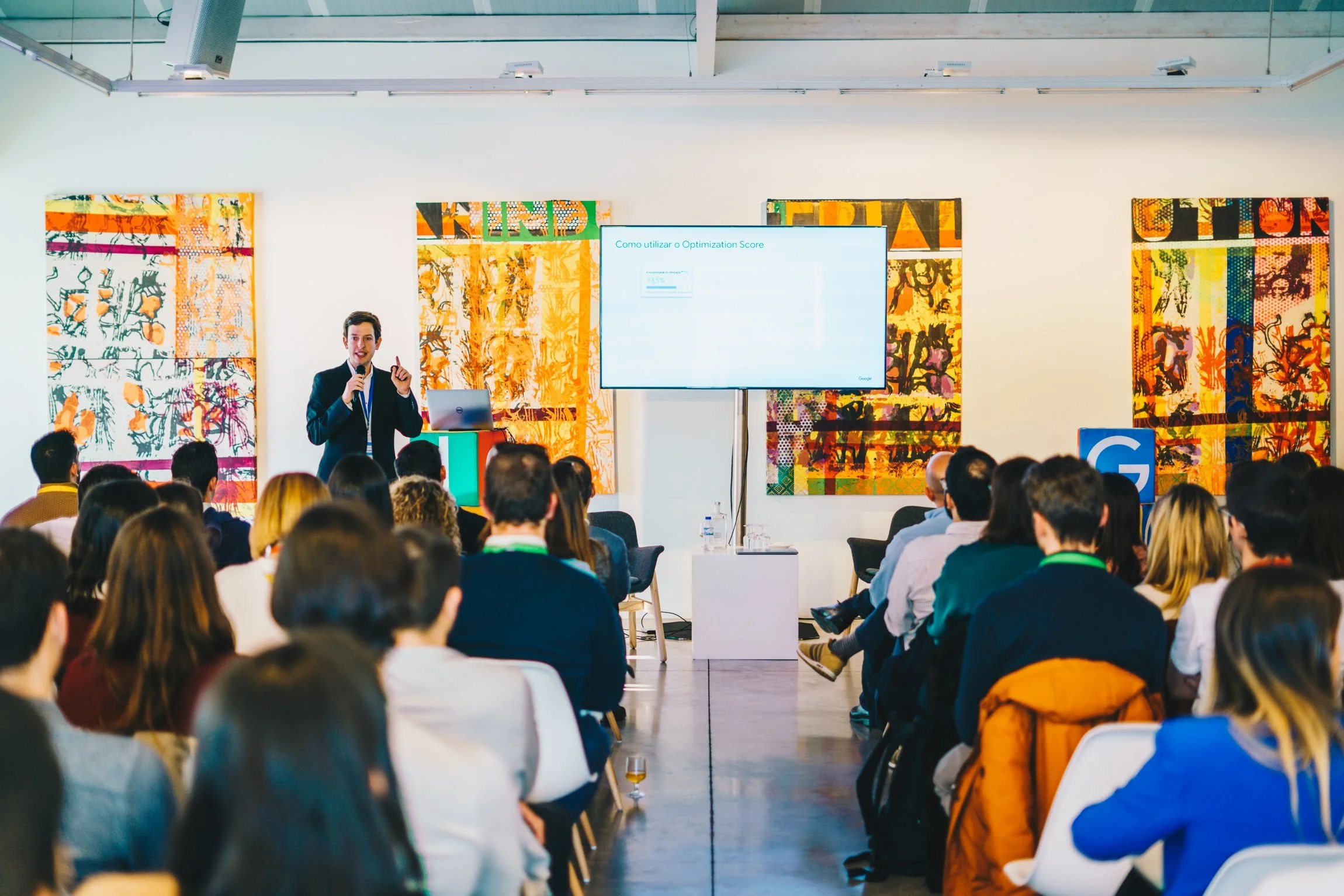A man in a suit giving a presentation to an audience in a modern conference room, with colorful abstract artwork on the walls and a large screen displaying information.