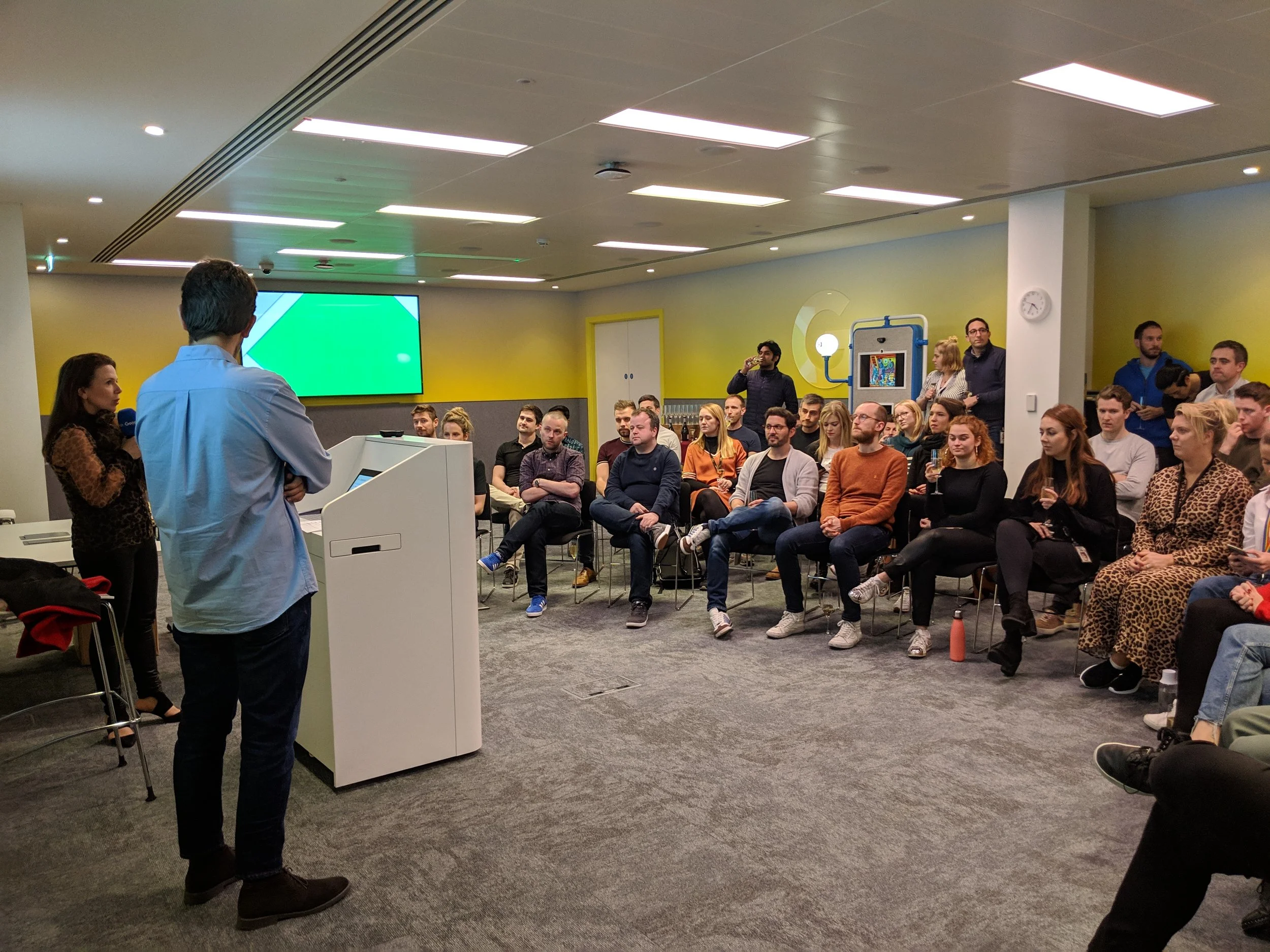 A man in a light blue shirt gives a presentation to an audience in a modern conference room, with a woman standing nearby. The audience, seated in rows, listens attentively.