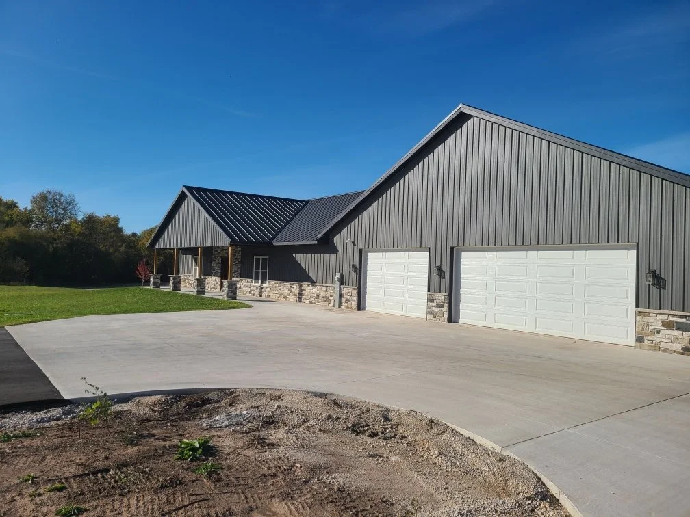 black standing seam steel roof on a black board and batten steel sided house with stone wainscot accents and white garage doors with a curved concrete driveway