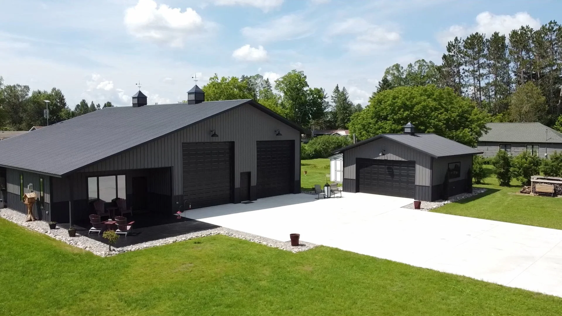 barndominium and shed with grey metal siding and black metal roofing and overhead garage doors with white long driveway in grassy neighborhood in Wisconsin