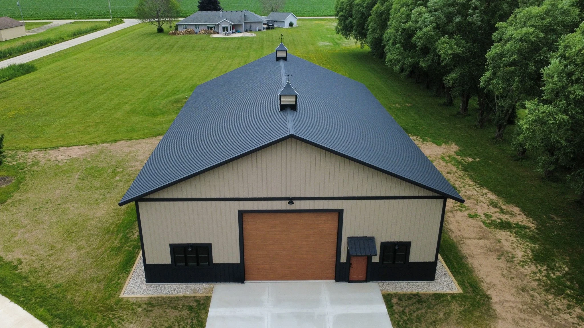 Clay textured steel siding and black textured steel roofing on post frame shed with wooden garage and service door and two cupolas in grassy field