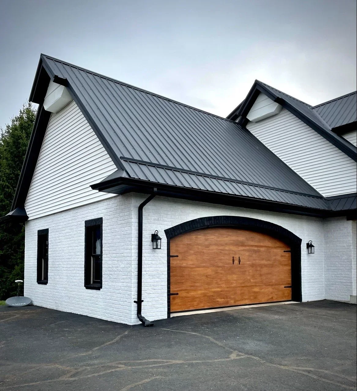 black standing seam metal roof on a white brick home with a wooden curved overhead garage door with black snow defenders on the roof