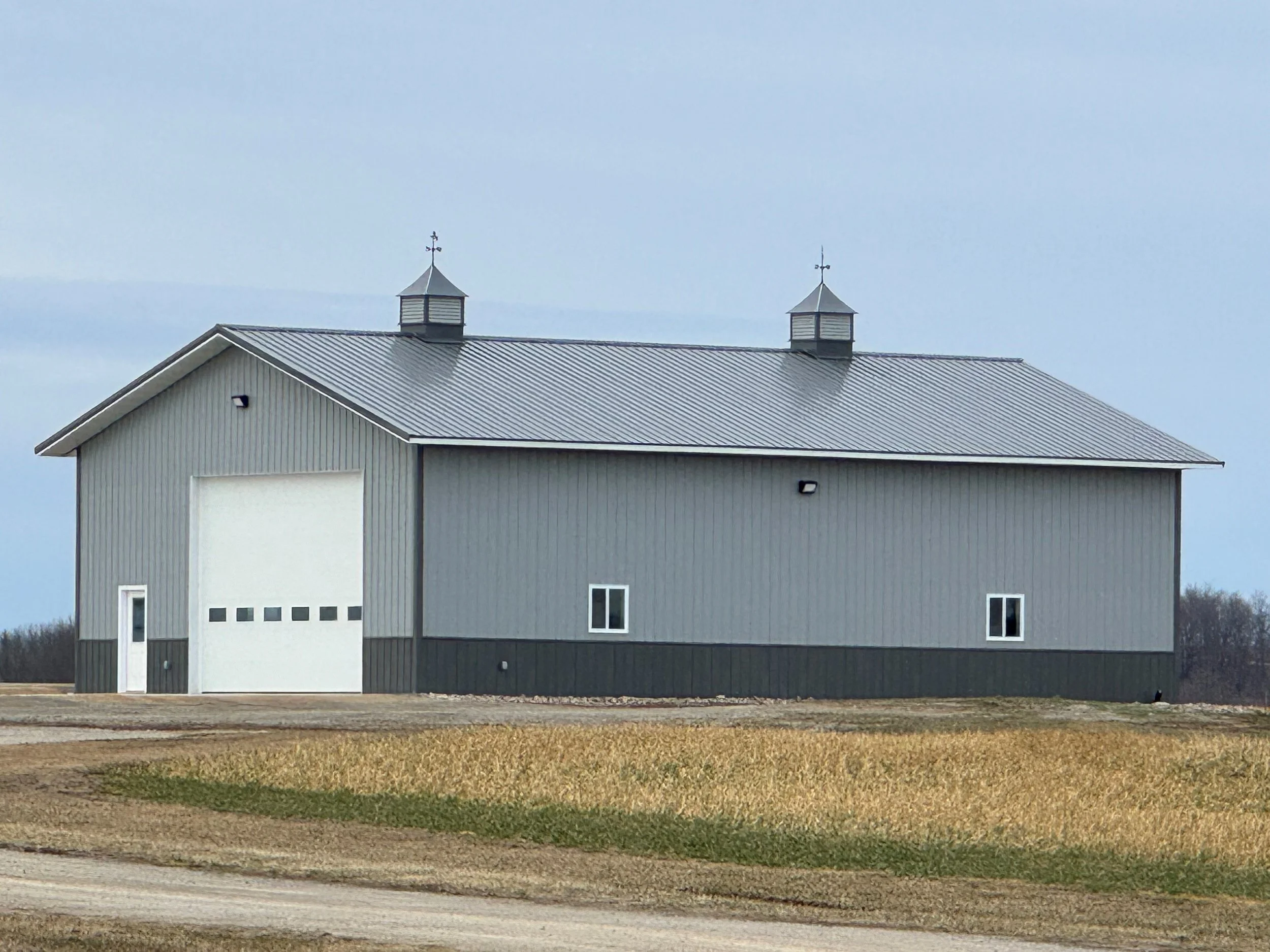 grey steel siding and dark grey steel roof and wainscot on post frame garage with double cupola in dead grassy field