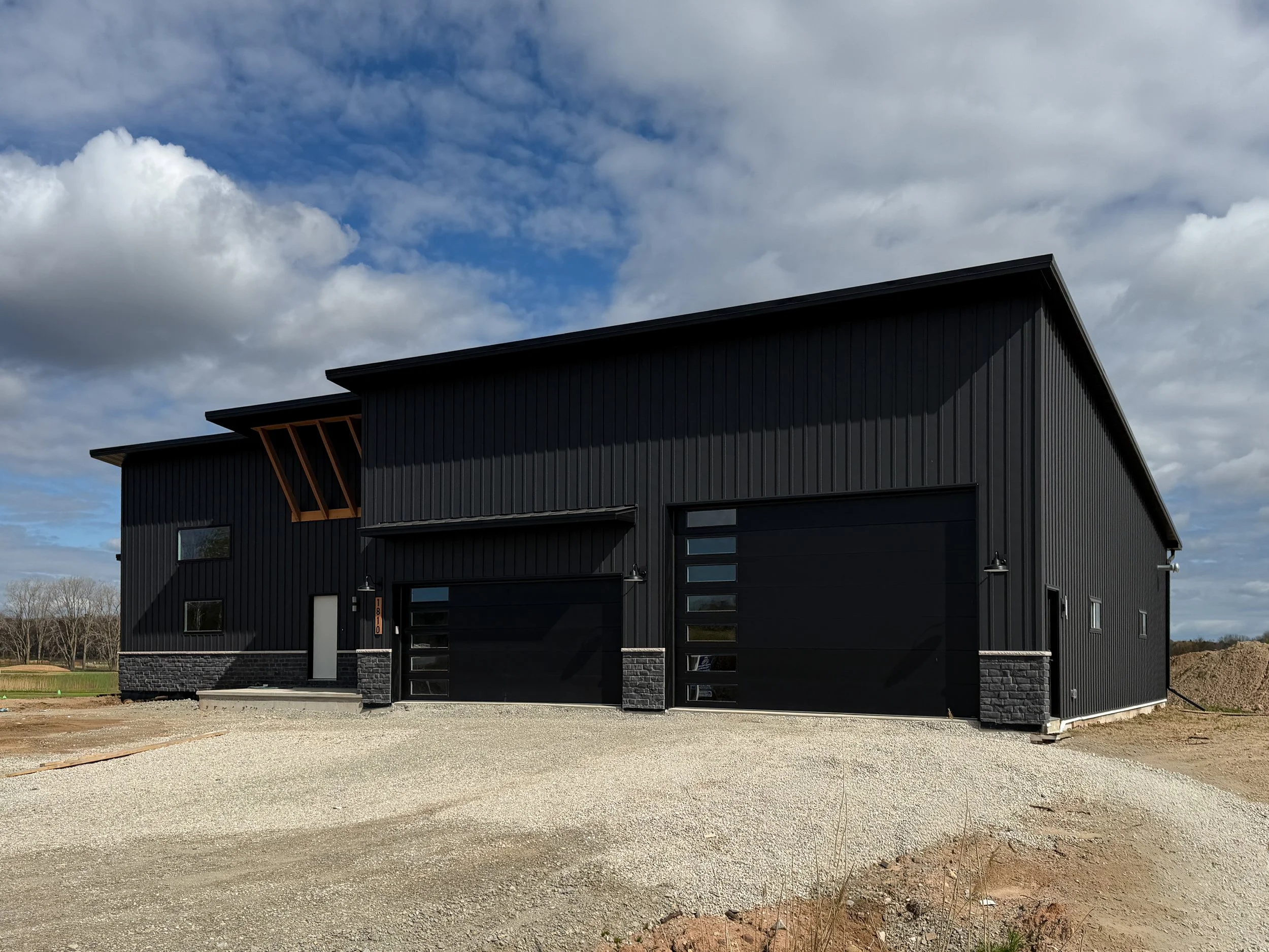 Modehome with black textured Board and Batten steel siding with black overhead garage doors, stone wainscot and wood accents and black standing seam roof